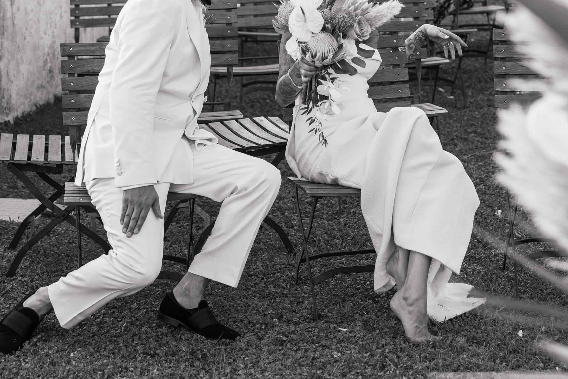 In bianco e nero, la sposa seduta tra le sedie della cerimonia e lo sposo che si piega verso di lei
