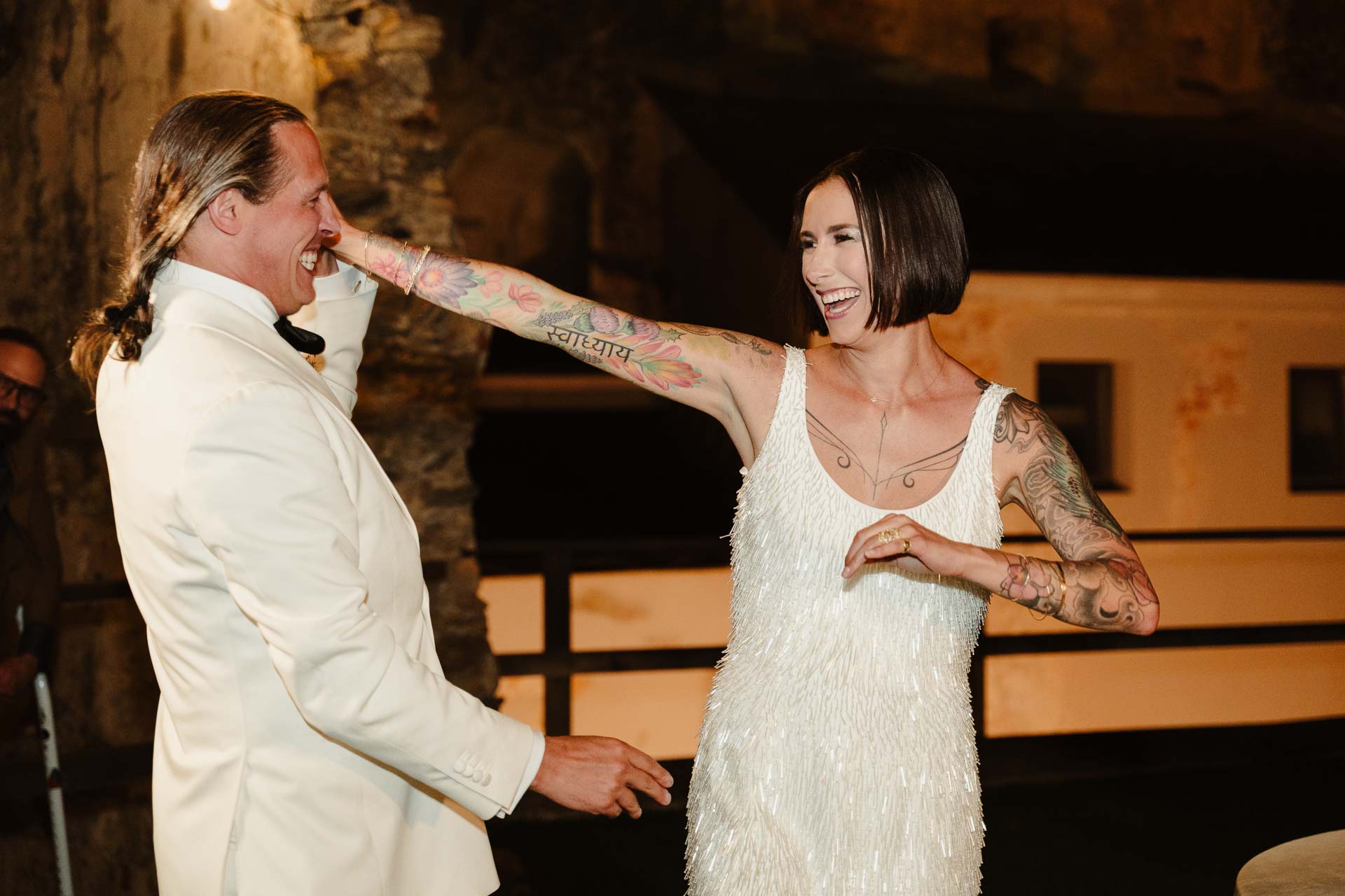 La sposa con il caschetto ride, il braccio tatuato teso verso lo sposo