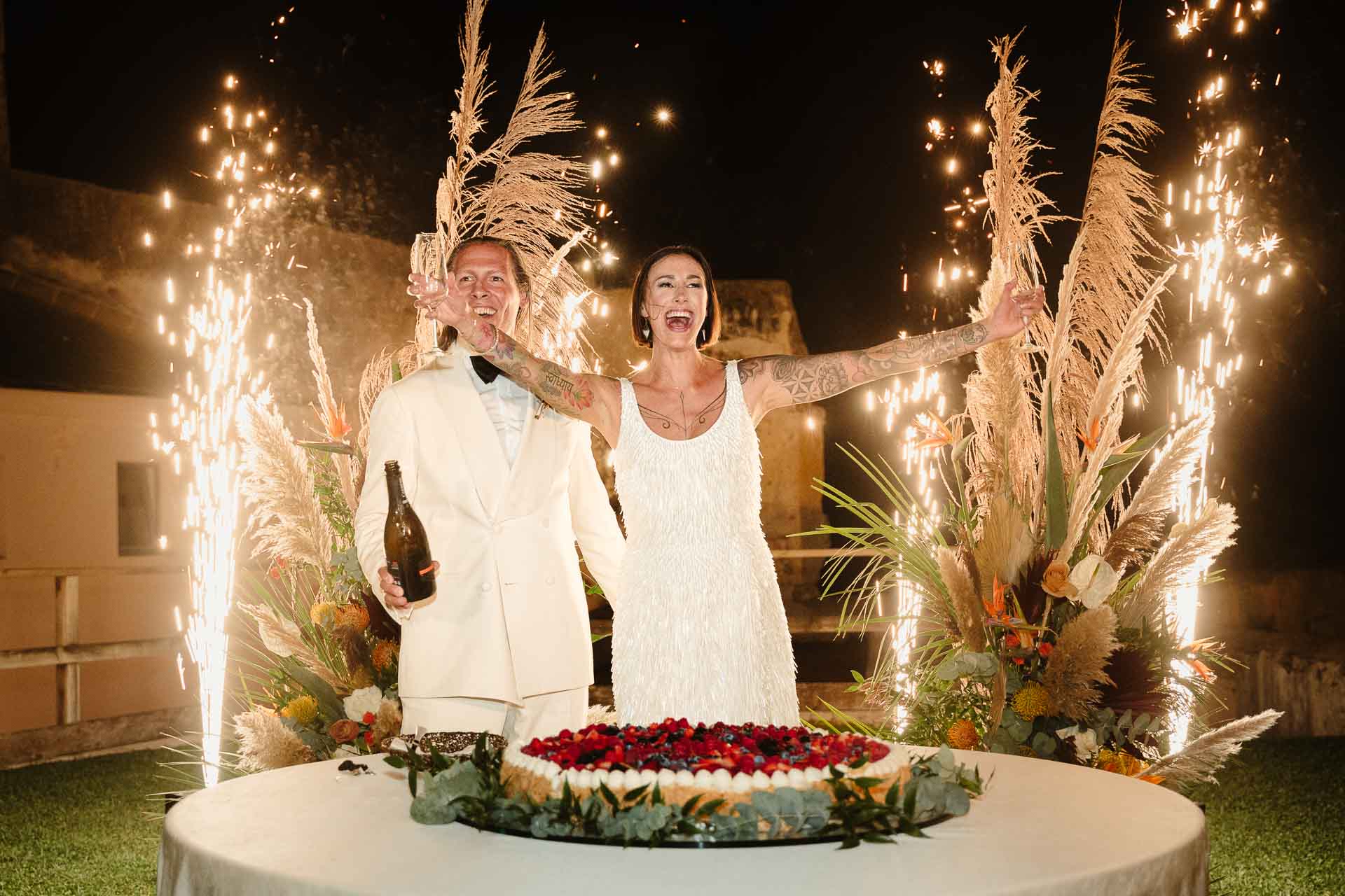 La sposa esulta a braccia aperte accanto allo sposo, dietro la torta tra le fontane di scintille