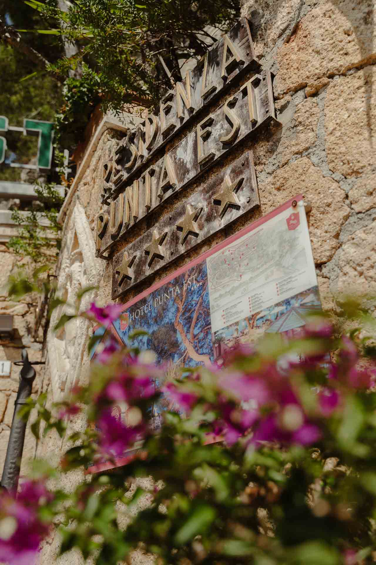 Un'insegna in ferro battuto sul muro di pietra, tra i fiori di buganvillea