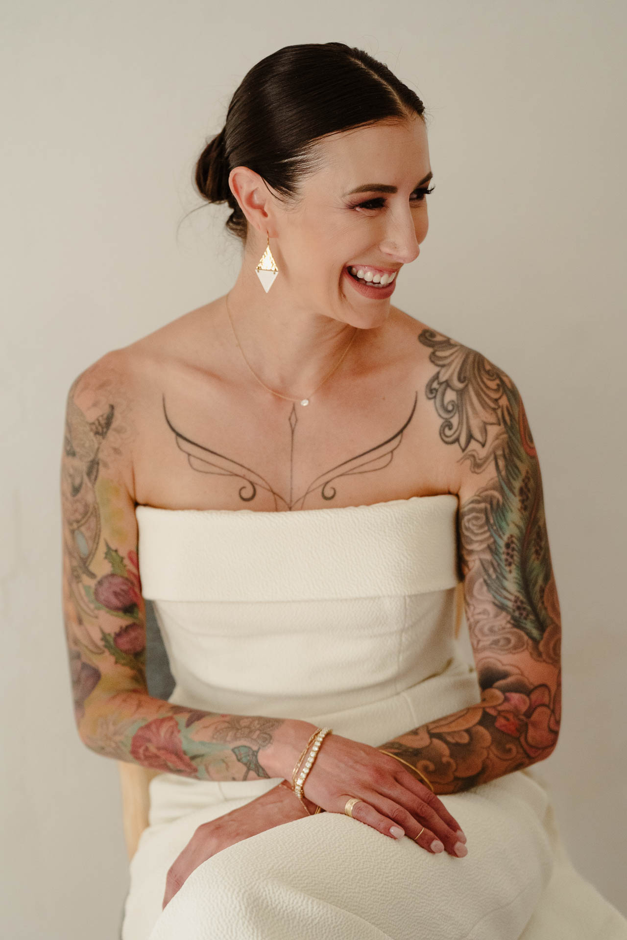 La sposa sorride seduta, con le braccia tatuate e gli orecchini a triangolo