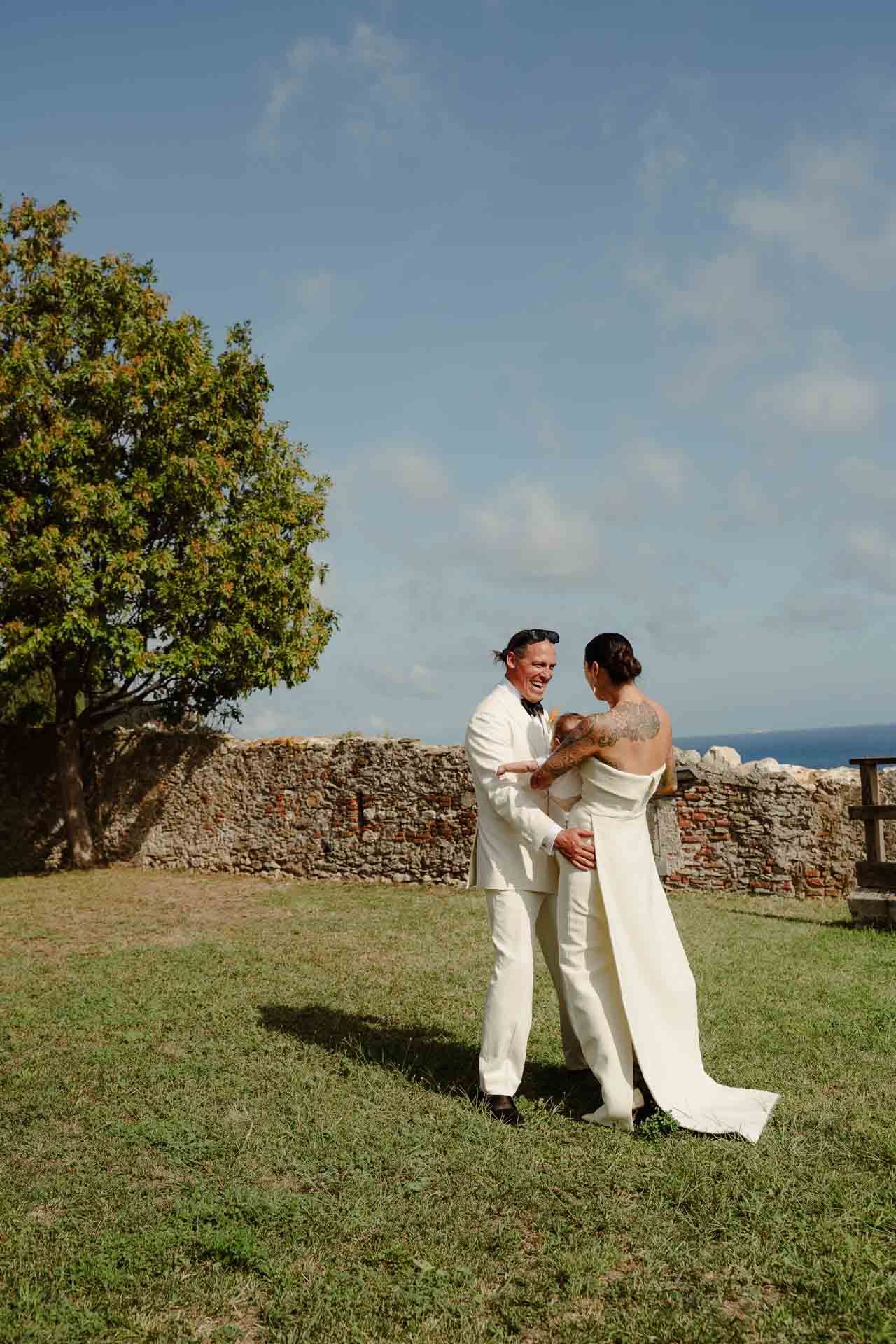 La sposa abbraccia lo sposo da dietro sul prato, sotto il cielo azzurro