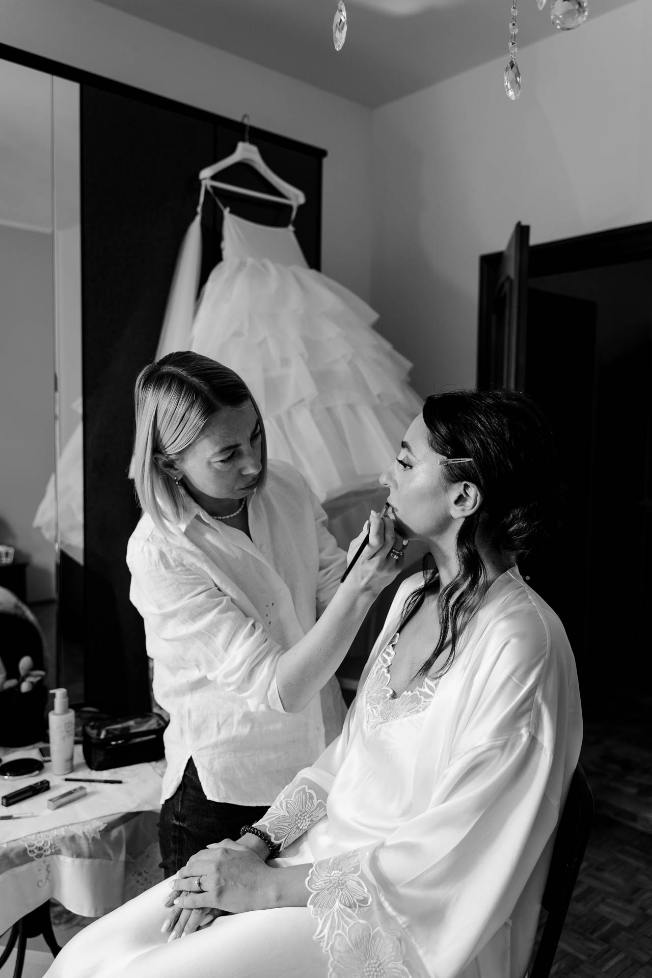 In bianco e nero, la truccatrice trucca la sposa seduta in vestaglia, con l'abito appeso alle spalle