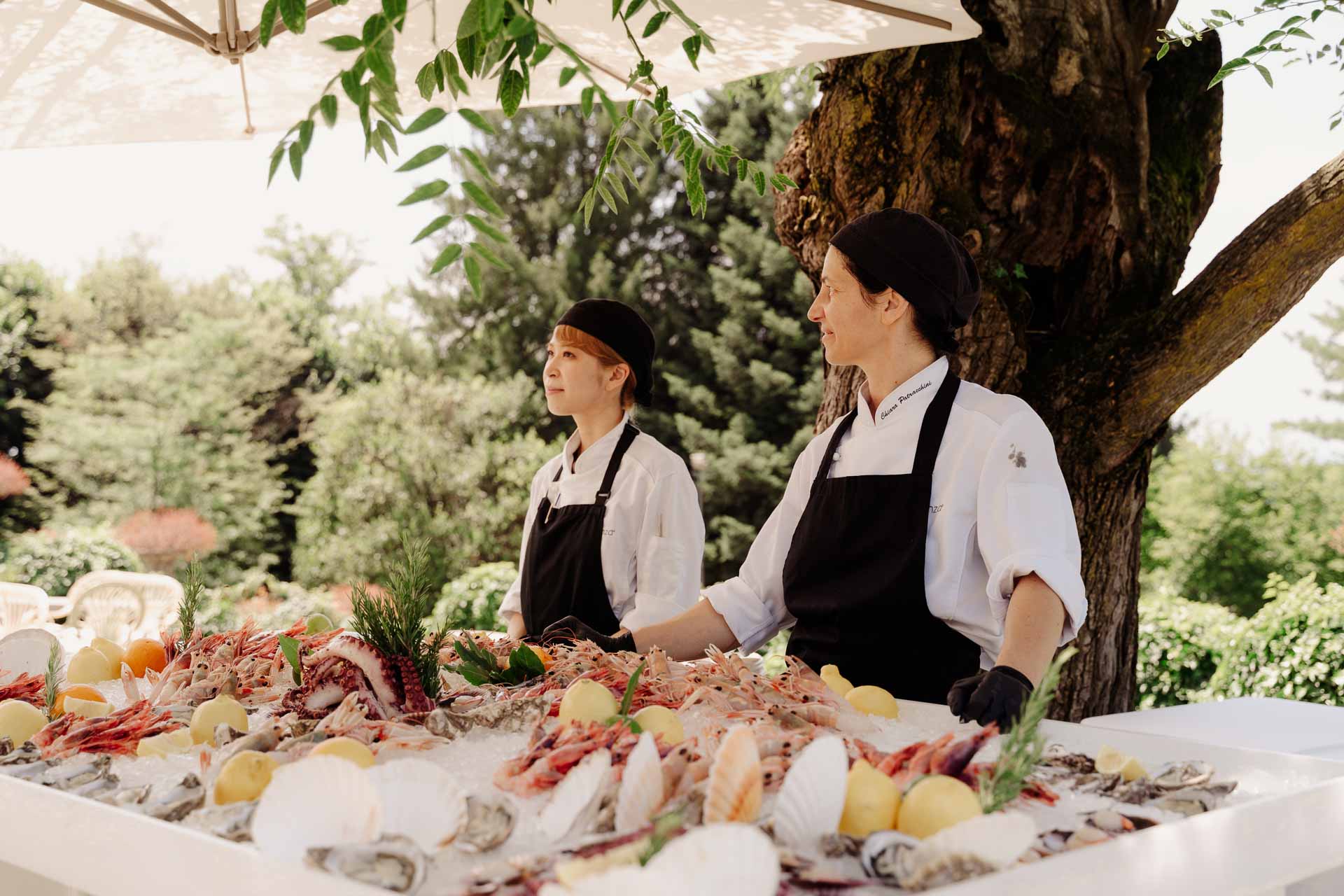 Due cuochi dietro il banco del crudo di mare, sotto gli alberi del parco