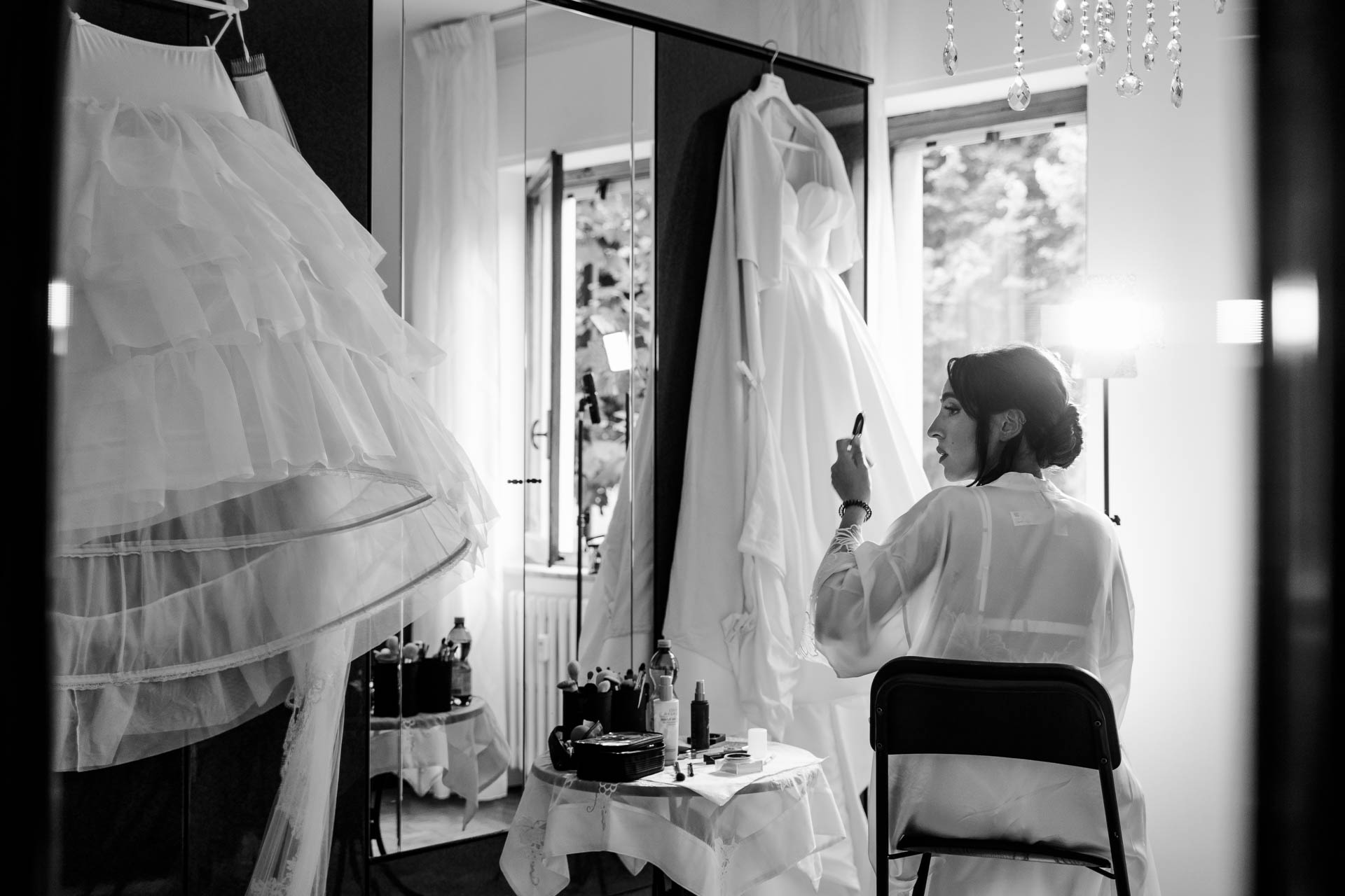 In bianco e nero, la sposa si trucca allo specchio nella stanza luminosa, tra il sottogonna e l'abito appeso
