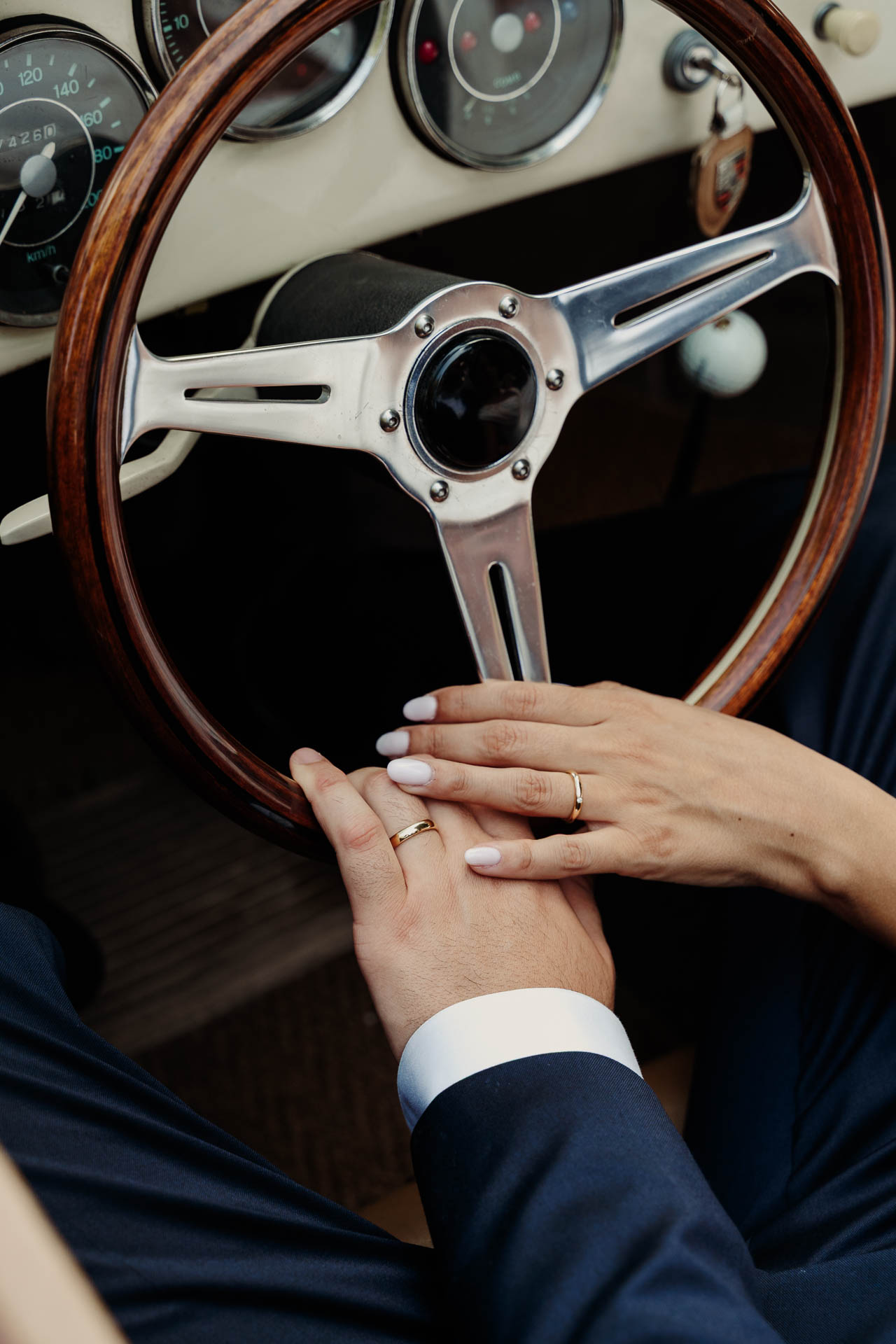 Le mani degli sposi con le fedi sul volante di legno dell'auto d'epoca