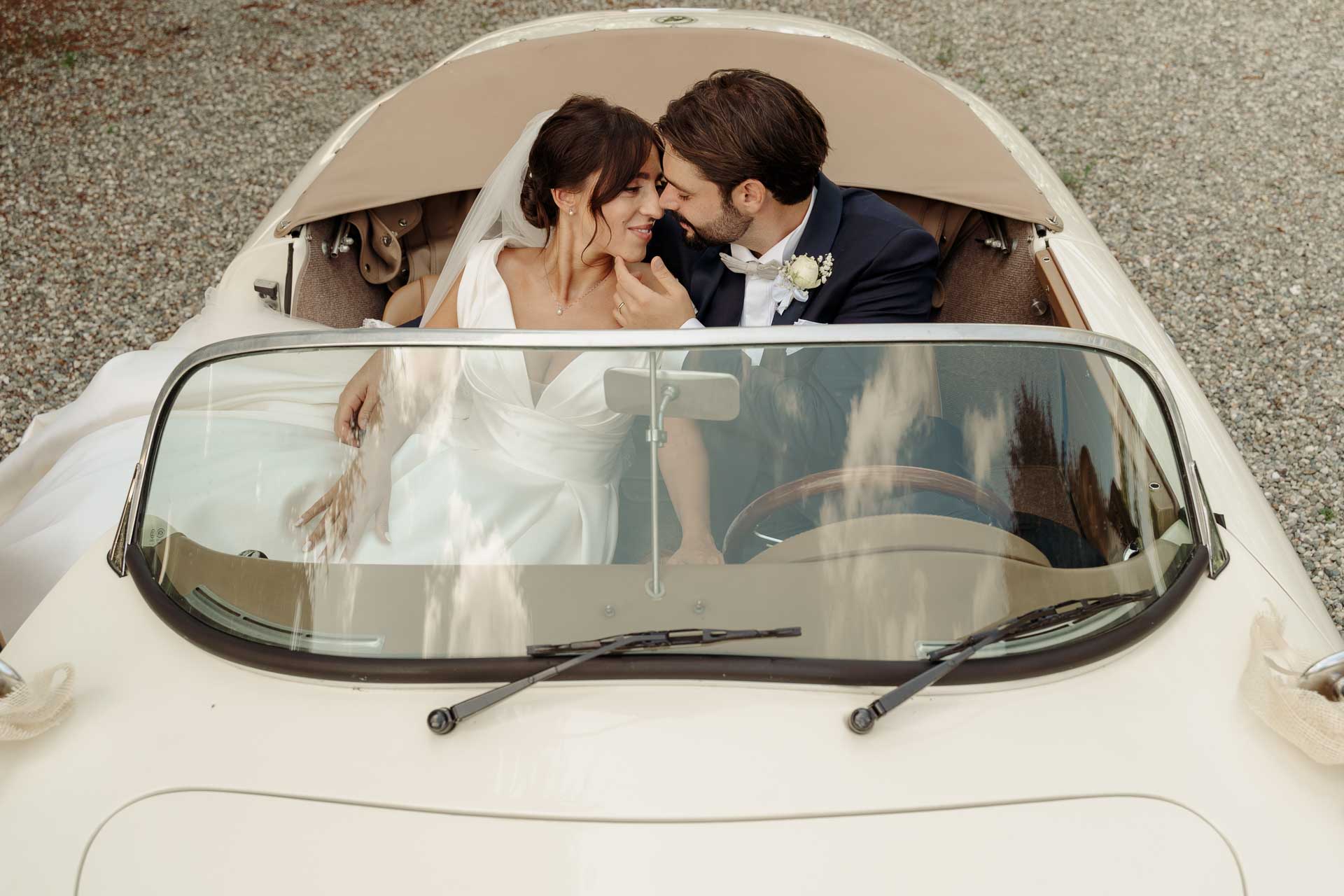 Gli sposi si guardano ridendo dentro l'auto d'epoca, visti da davanti