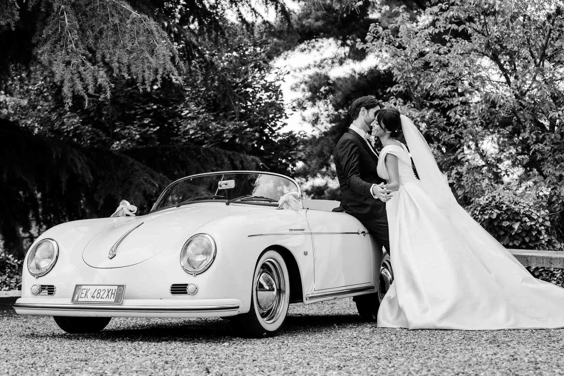 In bianco e nero, gli sposi si baciano davanti all'auto d'epoca, con lo strascico steso sulla ghiaia