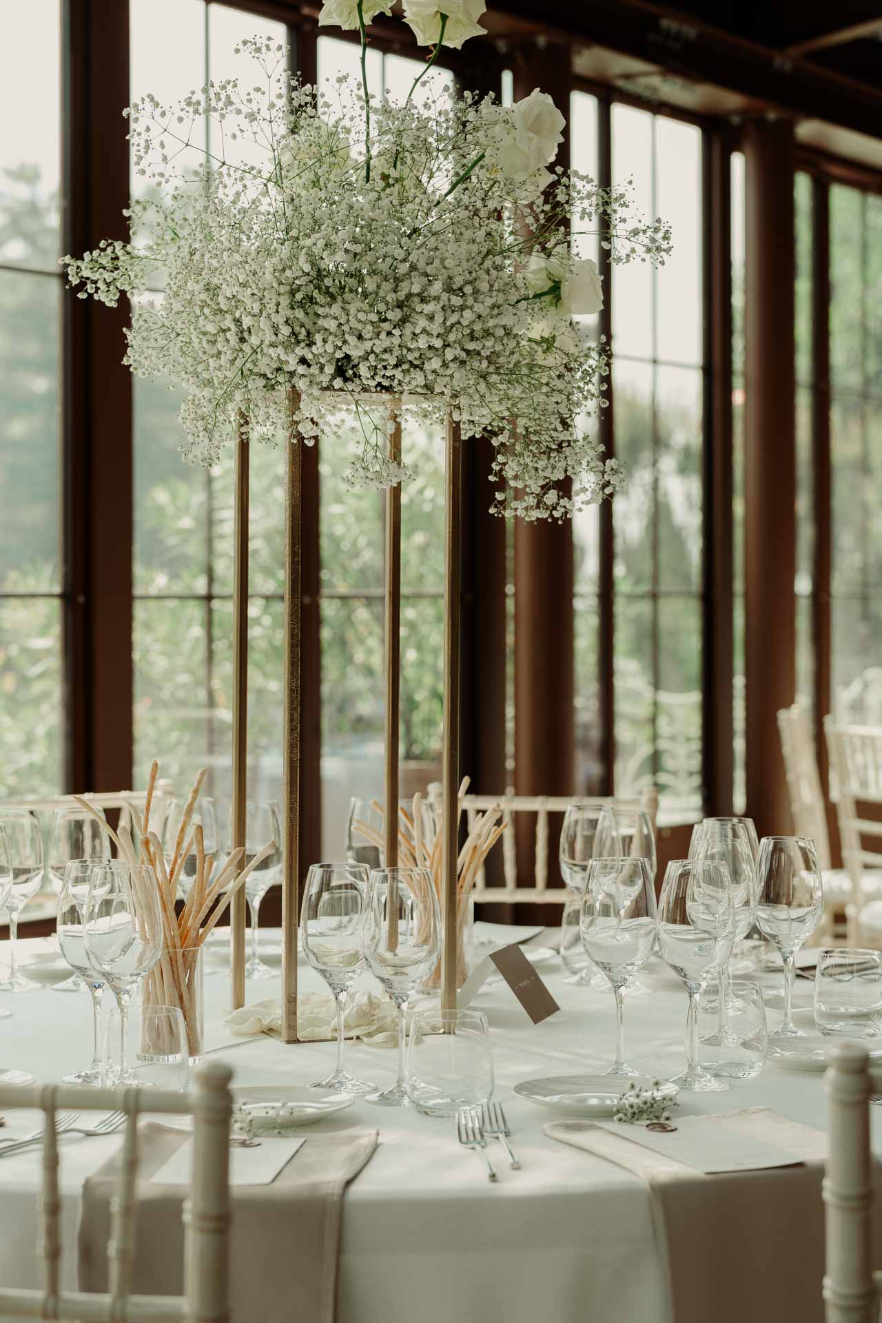 Un tavolo rotondo con una composizione alta di gypsophila e le spighe dorate, davanti alle vetrate