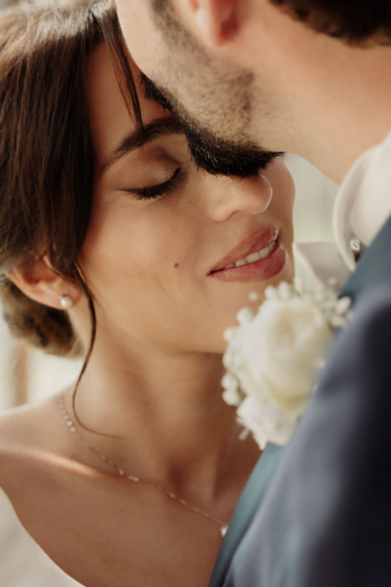 La sposa con gli occhi chiusi sorride contro la guancia dello sposo, con il fiore all'occhiello in primo piano