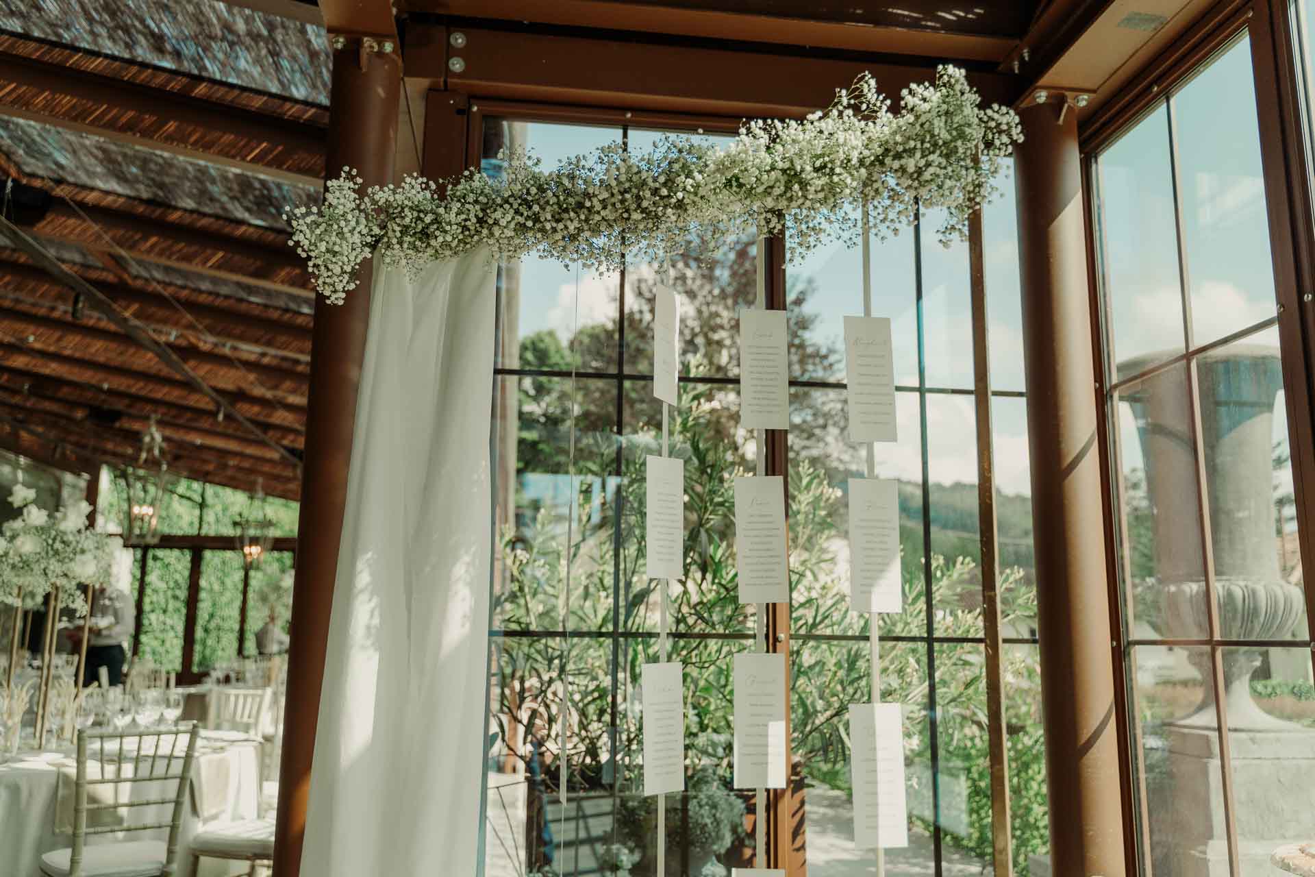 Il tableau de mariage appeso davanti alle vetrate, sotto un arco di gypsophila e un telo bianco