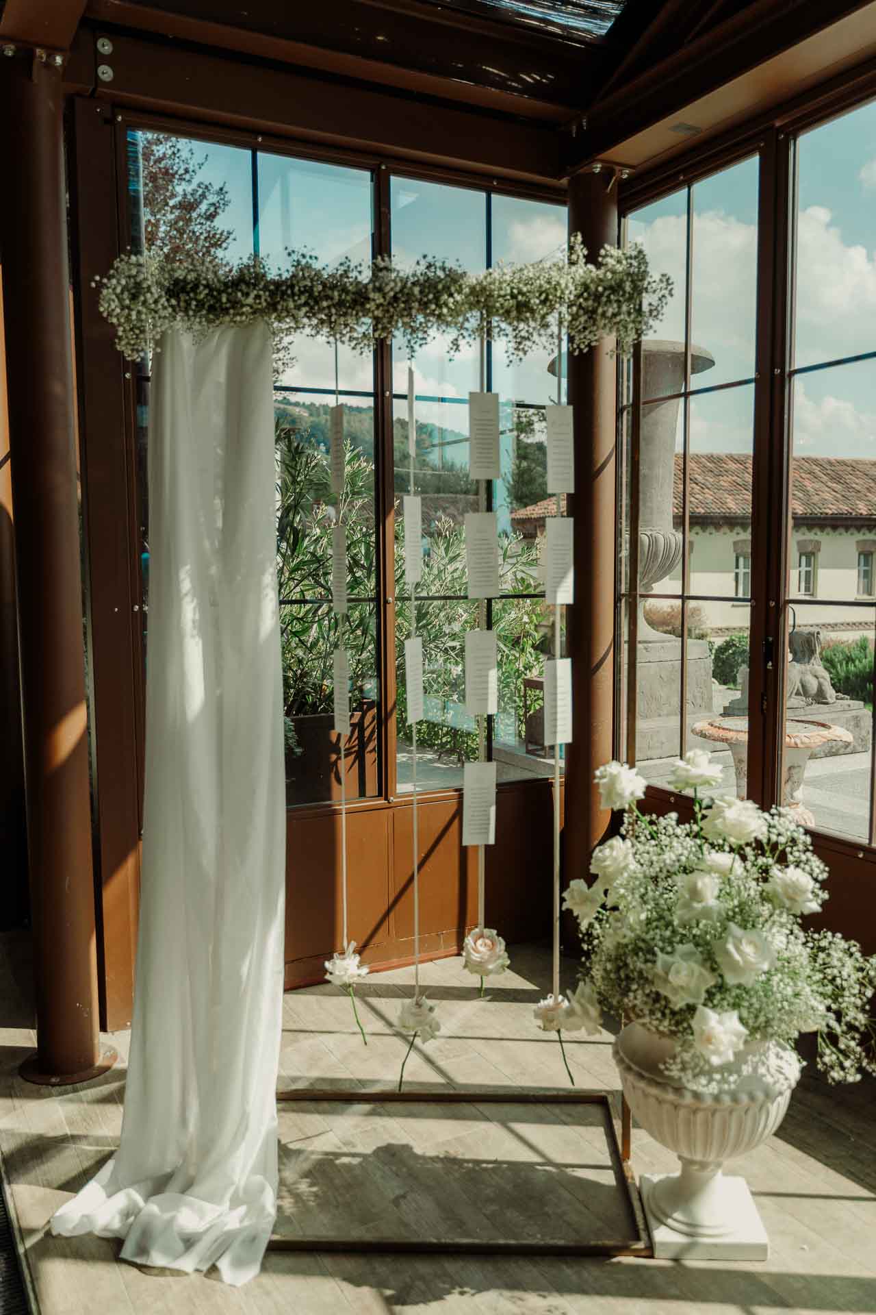 L'arco di gypsophila con il telo bianco davanti alle vetrate, e una coppa di fiori bianchi ai piedi