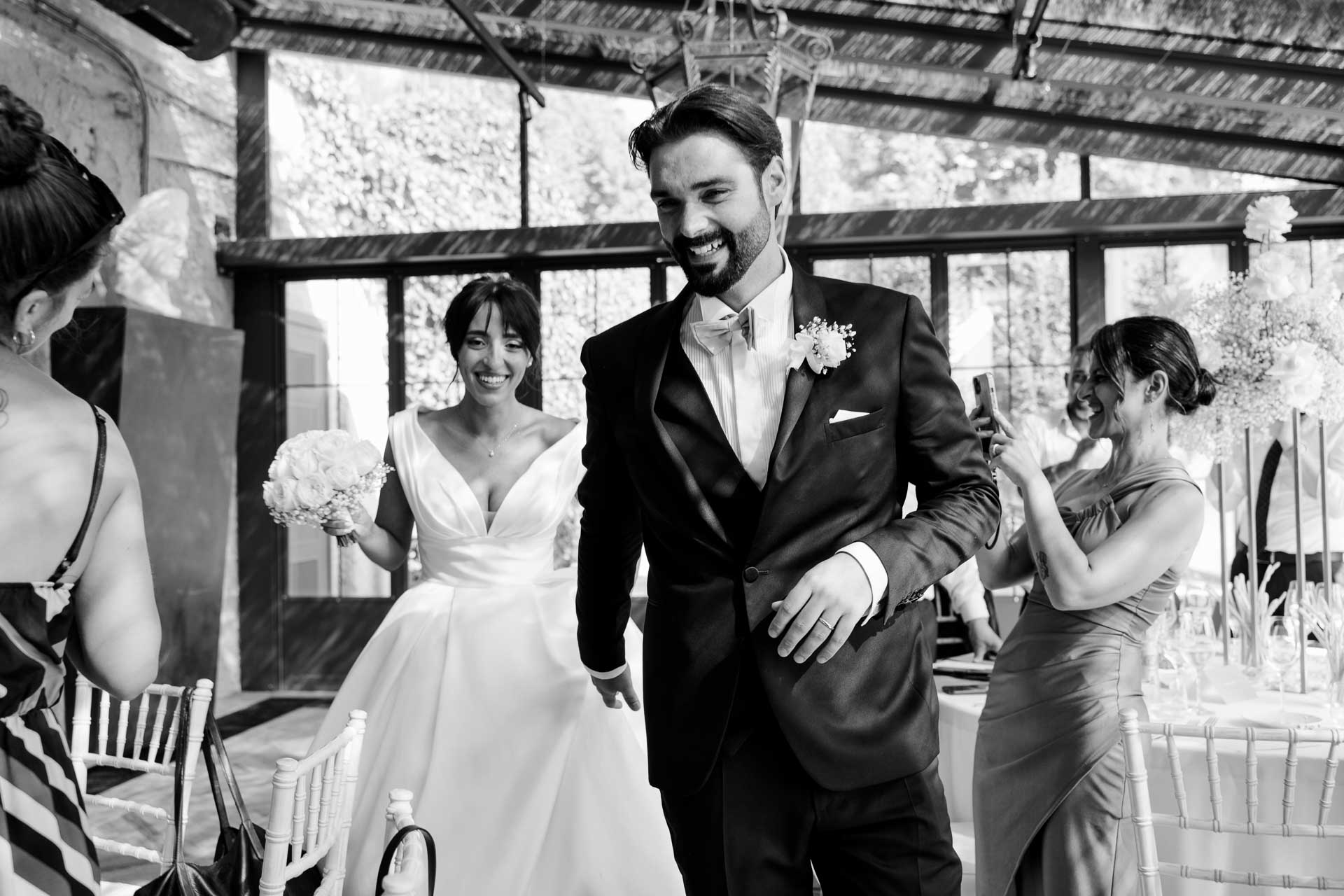 In bianco e nero, lo sposo ride entrando in sala accanto alla sposa, tra gli invitati che li fotografano
