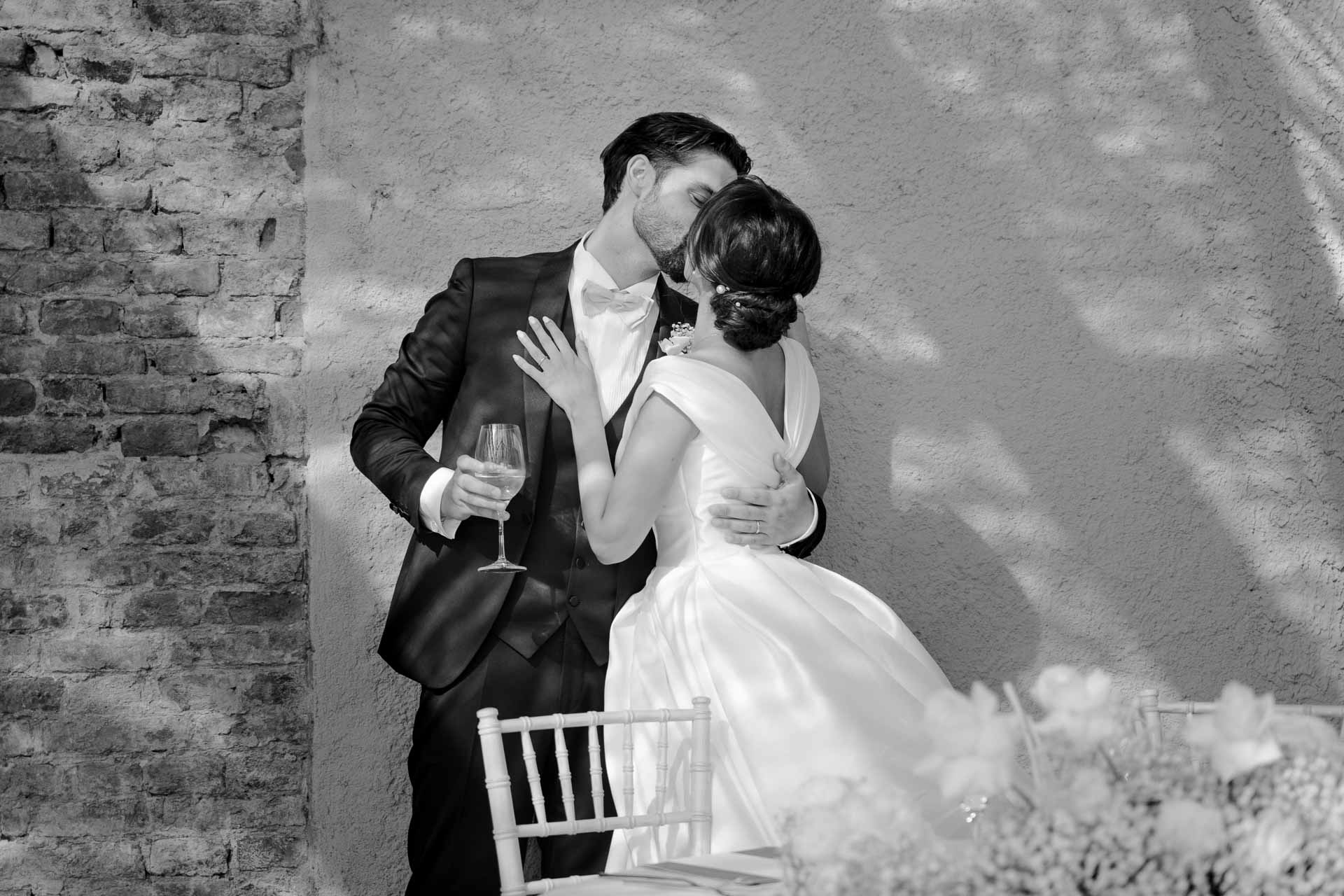 In bianco e nero, lo sposo con il calice bacia la sposa davanti al muro di mattoni