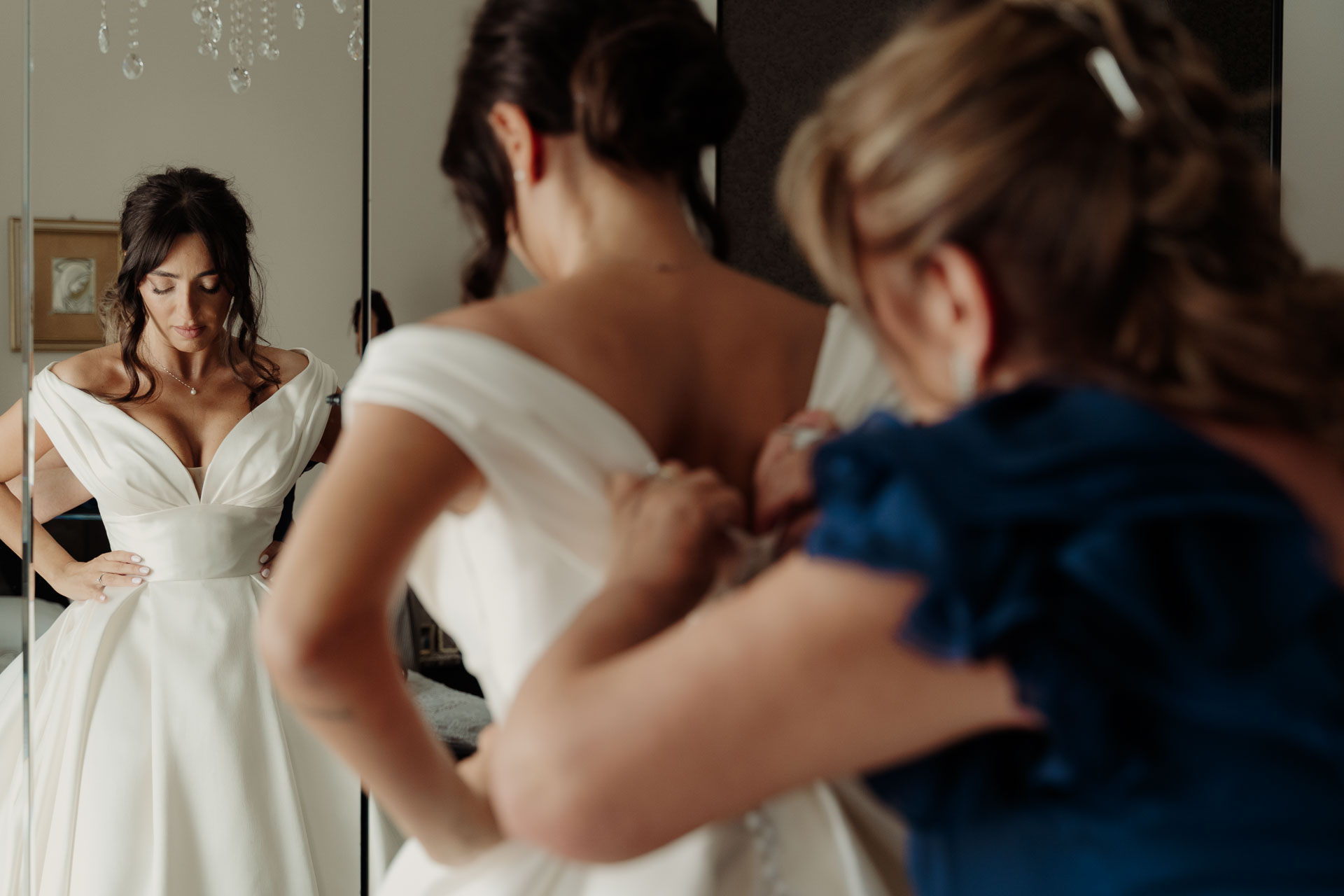 La sposa in abito davanti allo specchio, mentre una signora in blu le chiude i bottoni