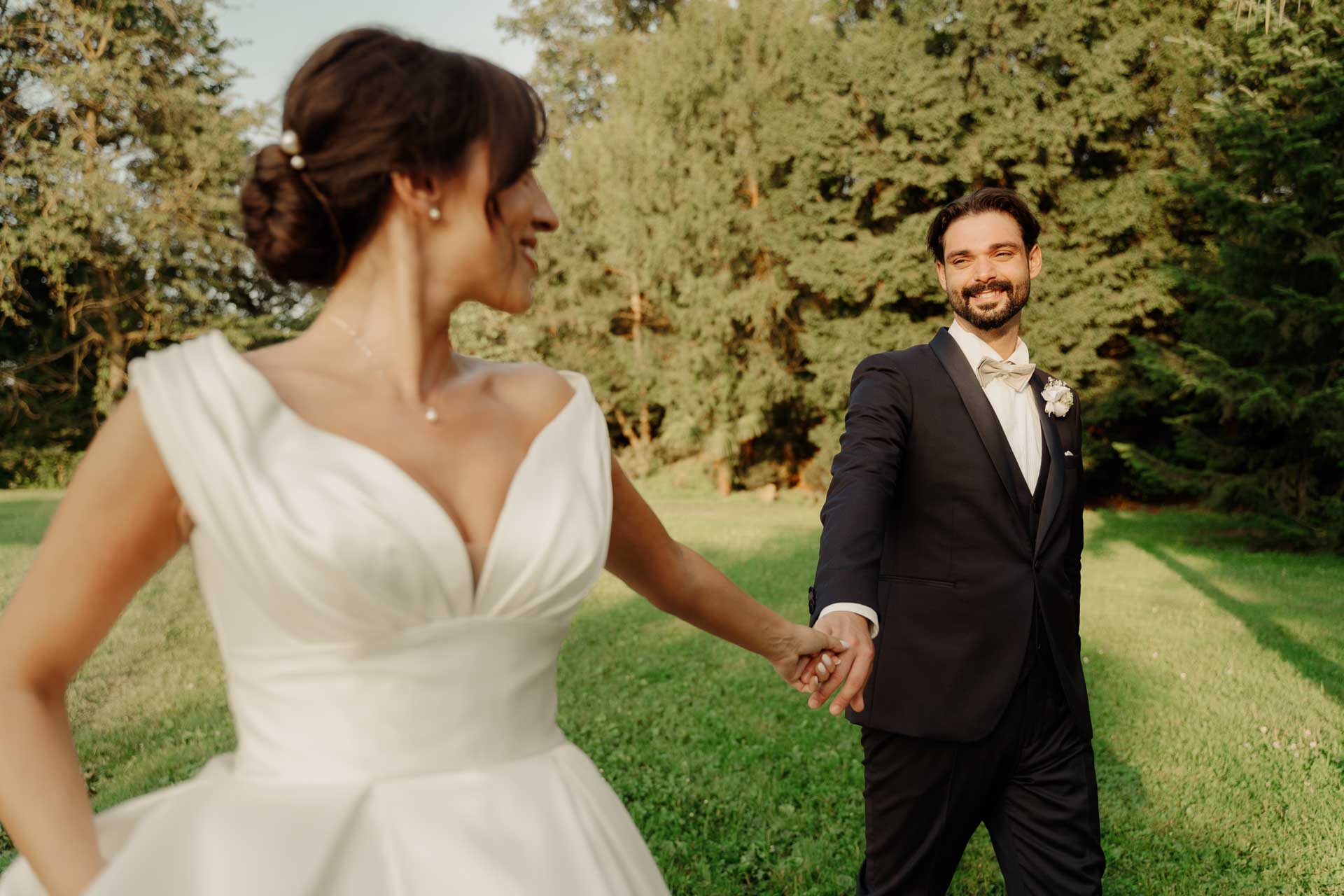 La sposa guarda lontano tenendo per mano lo sposo che sorride, sul prato al tramonto