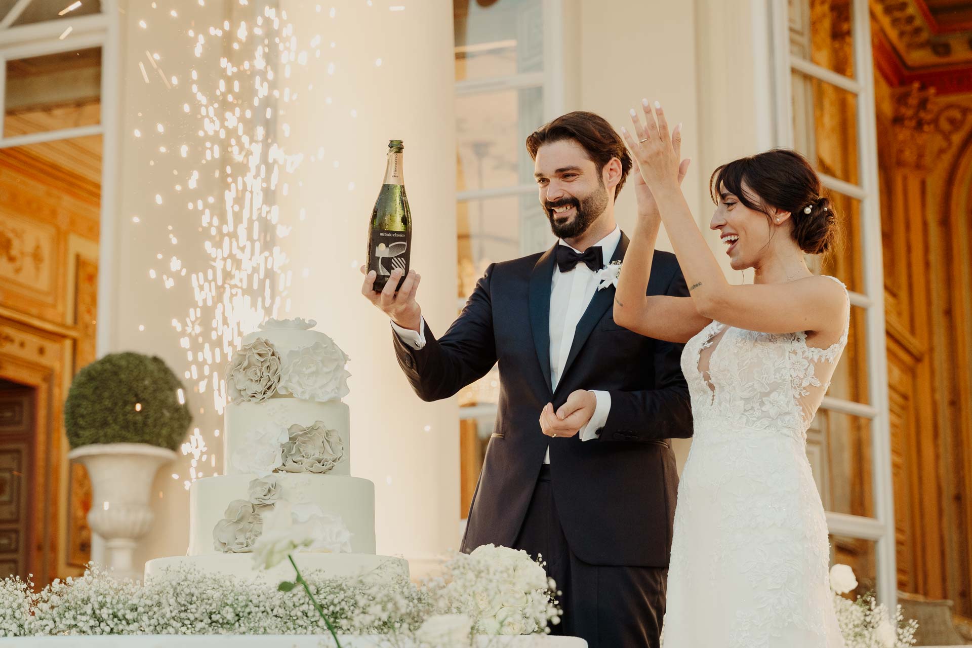 Lo sposo alza la bottiglia e la sposa applaude, davanti alla torta e alle fontane di scintille