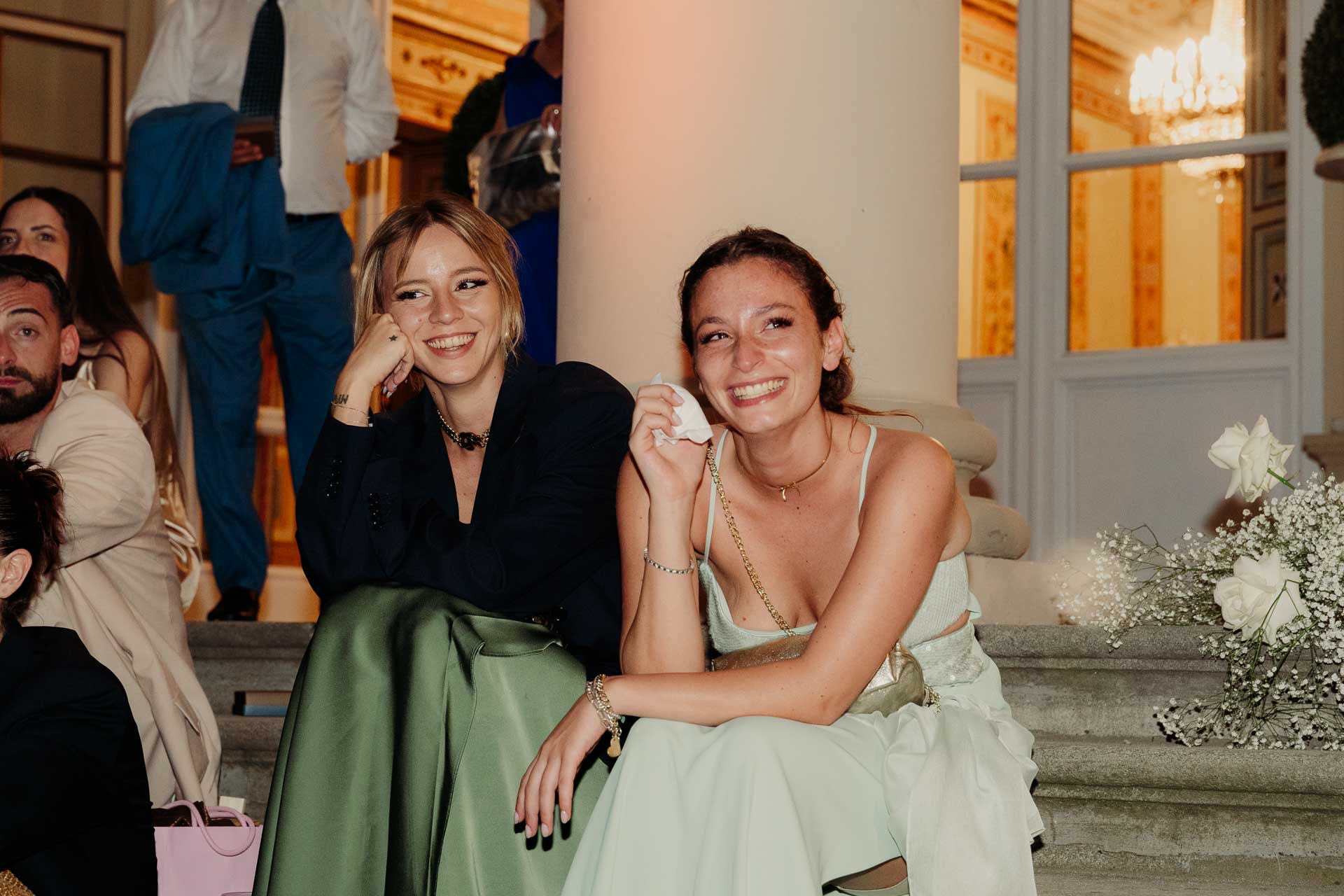 Due amiche sedute sui gradini ridono, una con un fazzoletto in mano