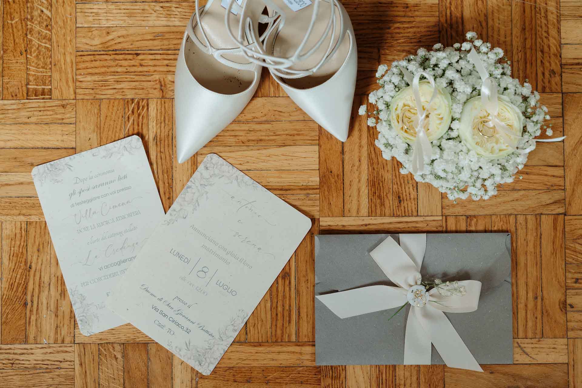 Le scarpe bianche, le partecipazioni e la busta grigia con il nastro, accanto a un cestino di gypsophila, sul parquet