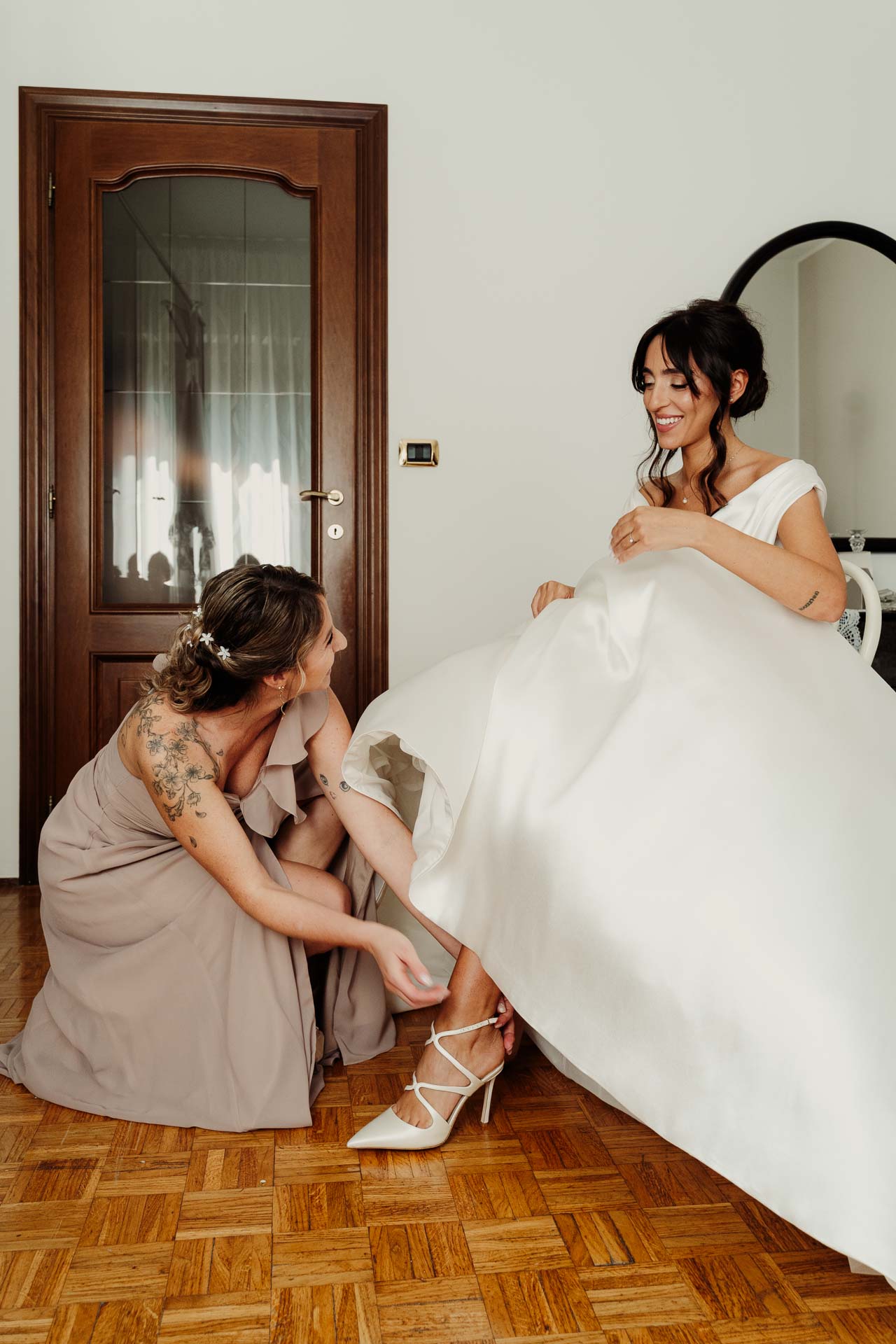 La sposa ride seduta con l'abito aperto sul parquet, mentre un'amica in rosa cipria, inginocchiata, le sistema la scarpa