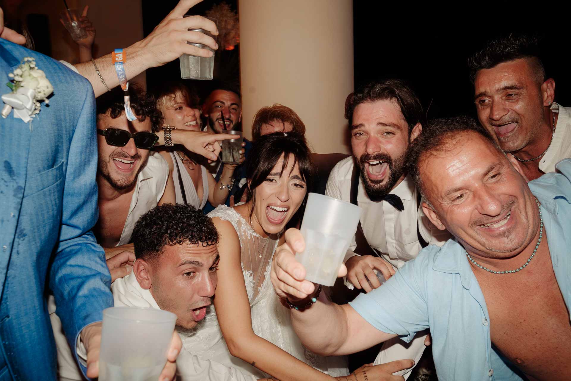 La sposa e lo sposo stretti dagli amici che ridono e brindano, a fine serata