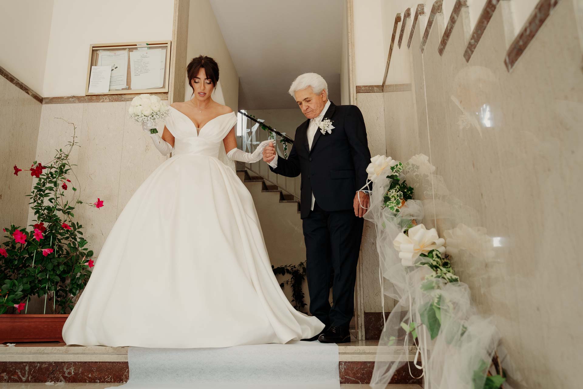 La sposa in abito scende le scale del portone sottobraccio a un signore in abito scuro, tra i fiori e i fiocchi