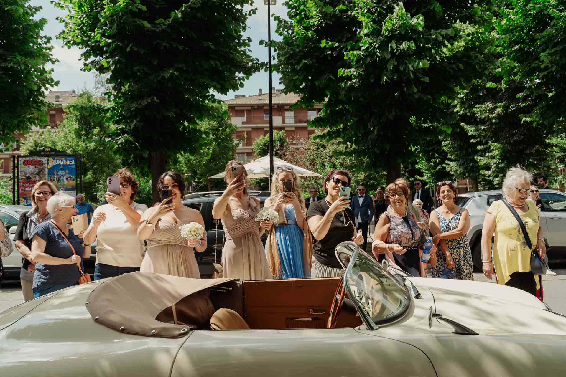 Una folla di parenti e vicini con i telefoni in mano aspetta la sposa davanti all'auto d'epoca bianca