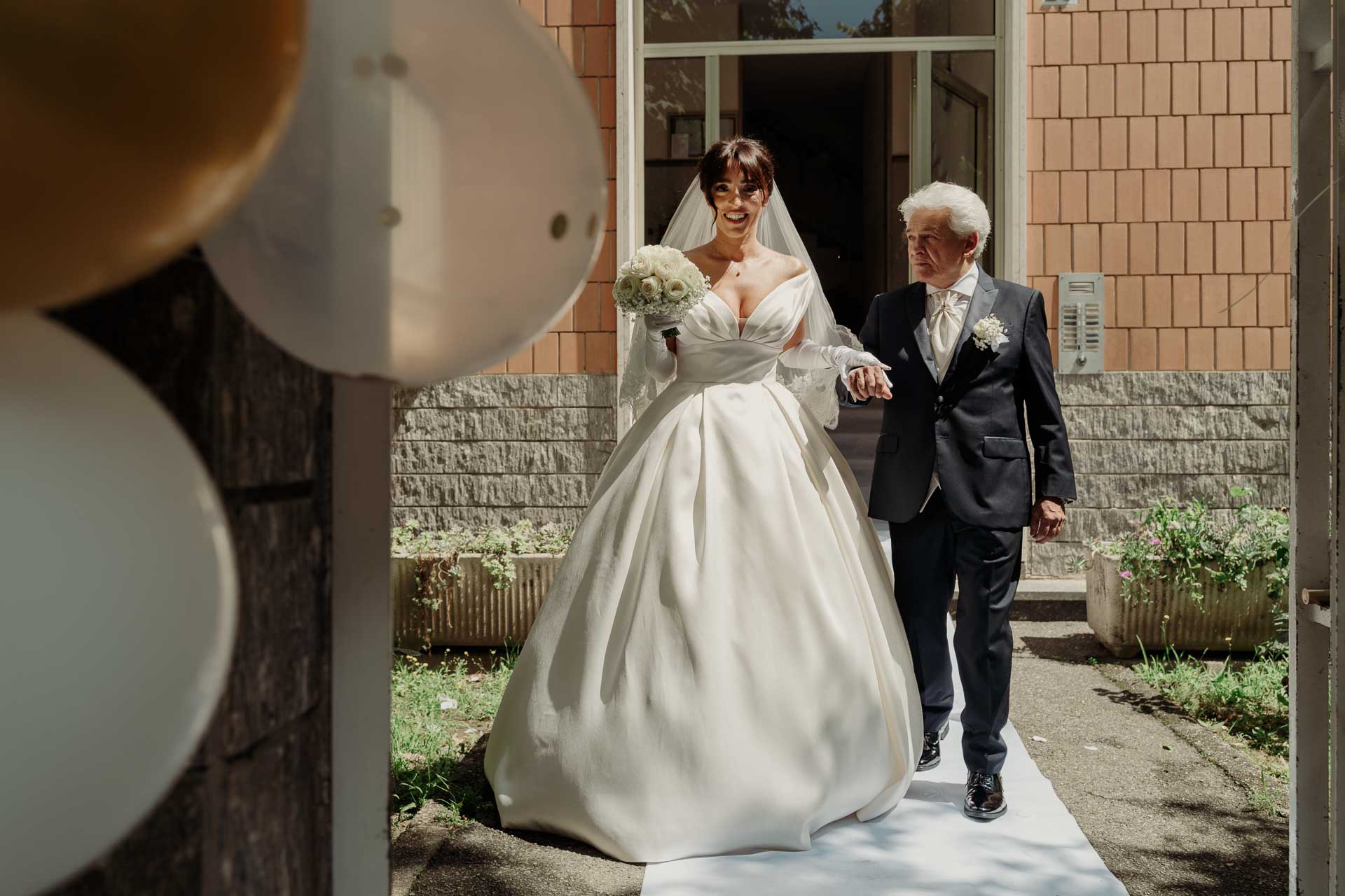 La sposa esce dal portone a braccio di un signore in abito scuro, tra i palloncini