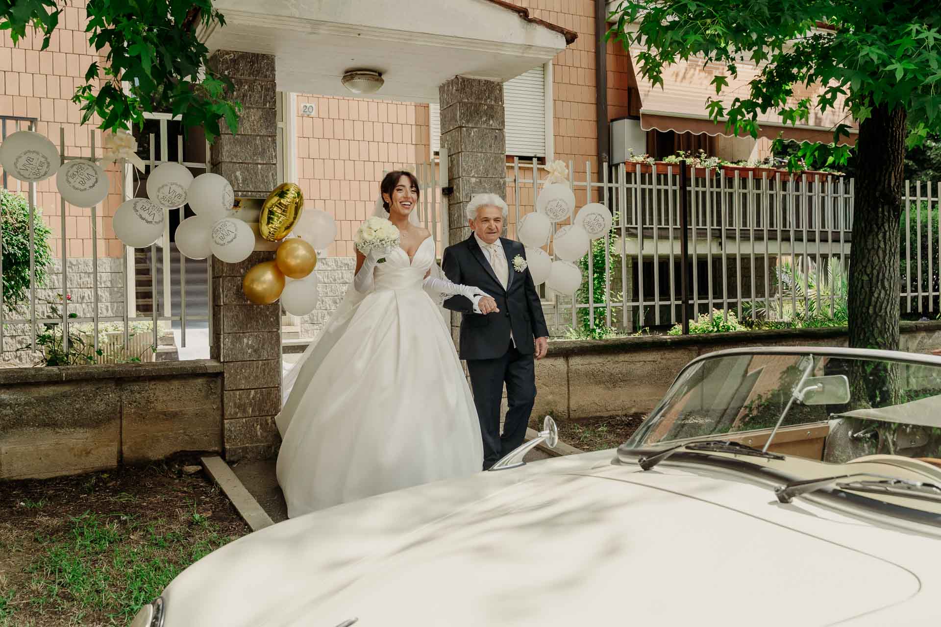 La sposa e un signore in abito scuro nel giardino davanti all'auto d'epoca bianca, tra i palloncini