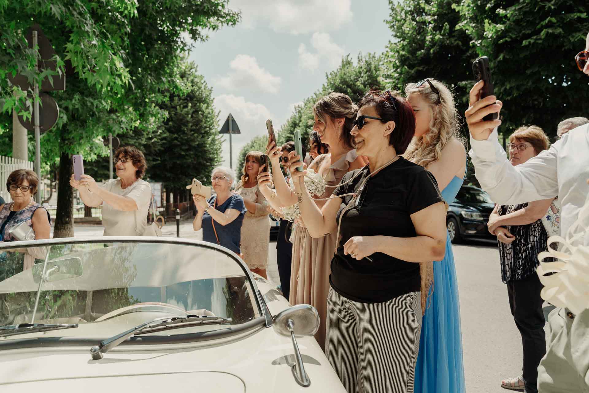 Le invitate fotografano la sposa con i telefoni, dietro il parabrezza dell'auto d'epoca