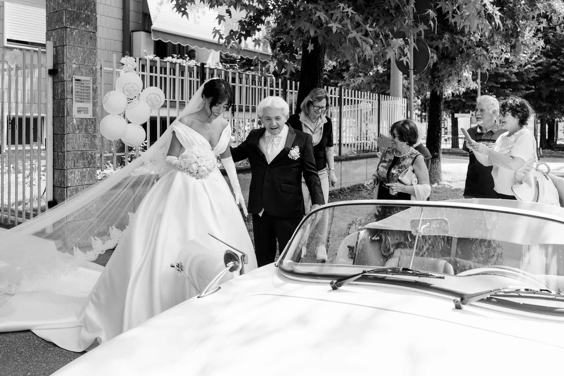 In bianco e nero, la sposa sale sull'auto d'epoca con il velo steso, aiutata da un signore in abito scuro