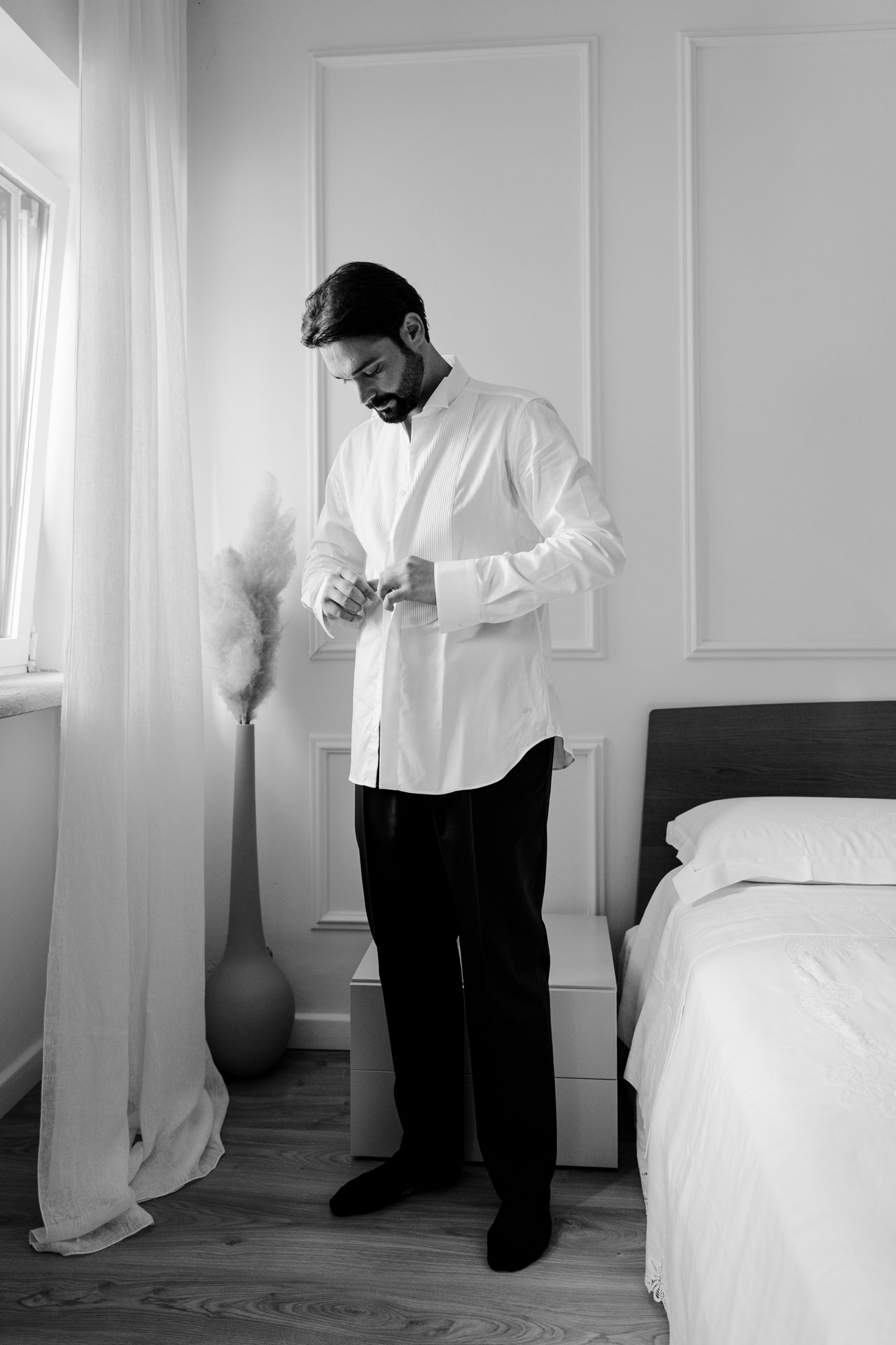 In bianco e nero, lo sposo in camicia bianca si abbottona i polsini accanto alla finestra