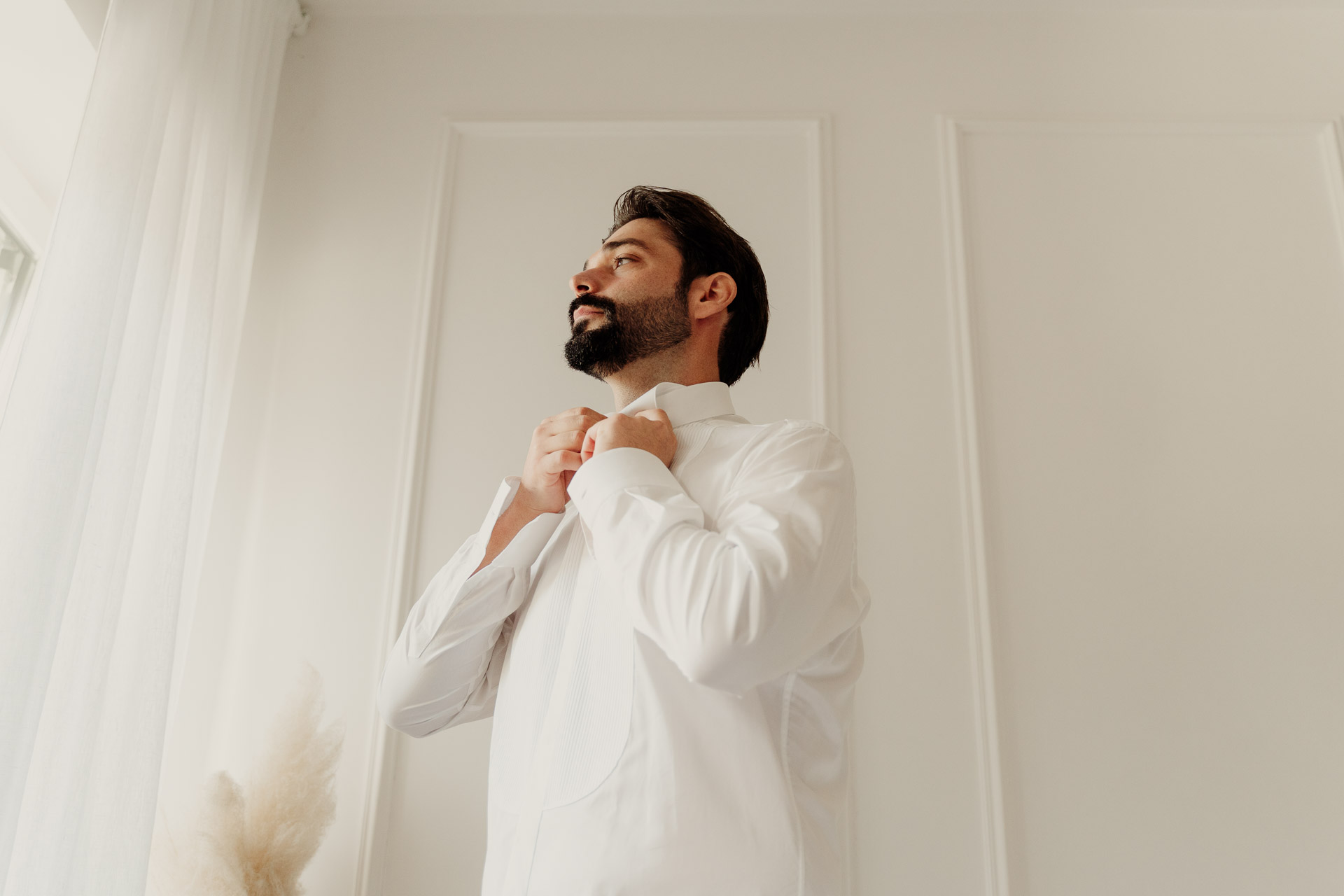 Lo sposo alza lo sguardo mentre si sistema il colletto della camicia davanti alla parete bianca