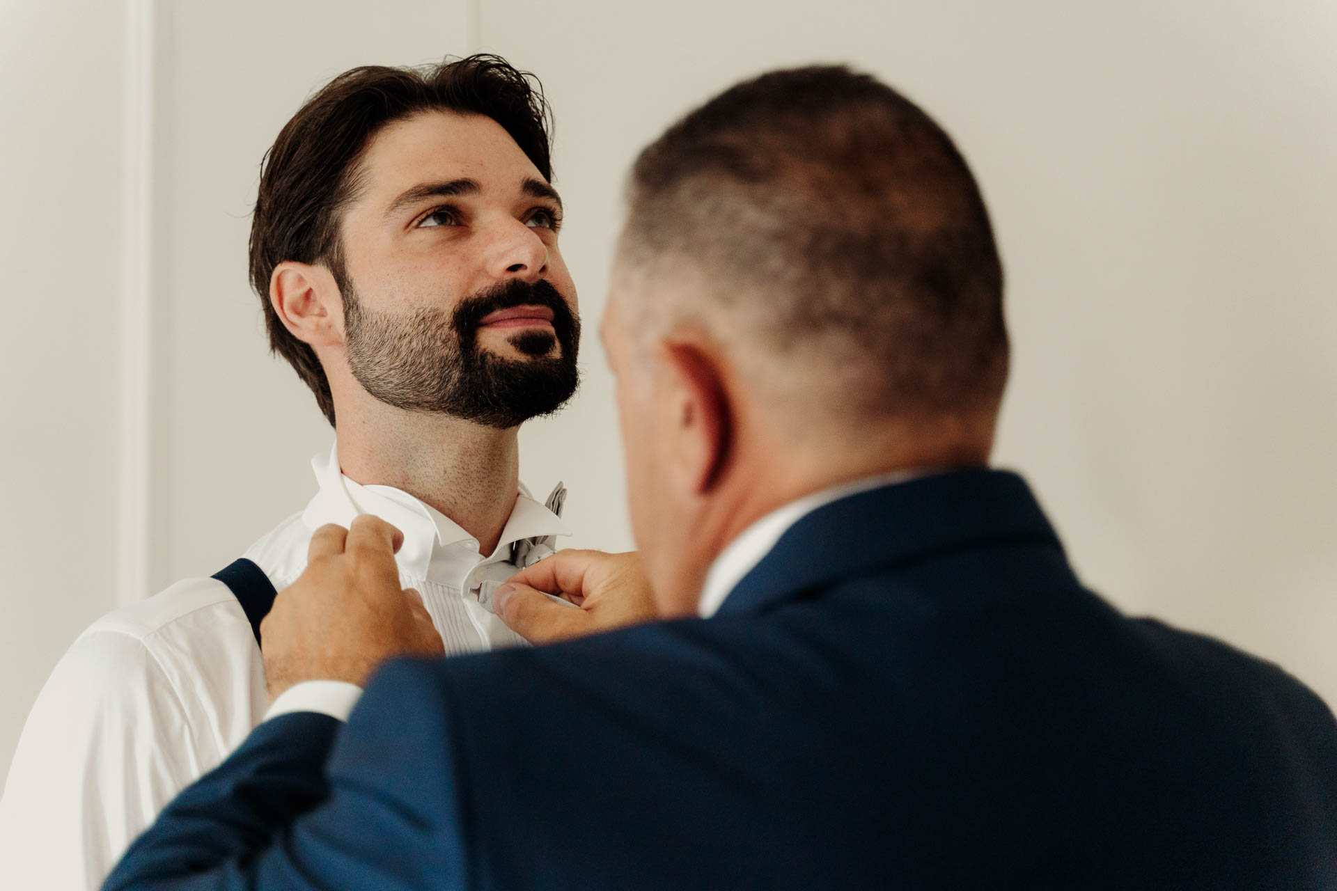 Un uomo in giacca blu sistema il papillon allo sposo, che guarda in alto