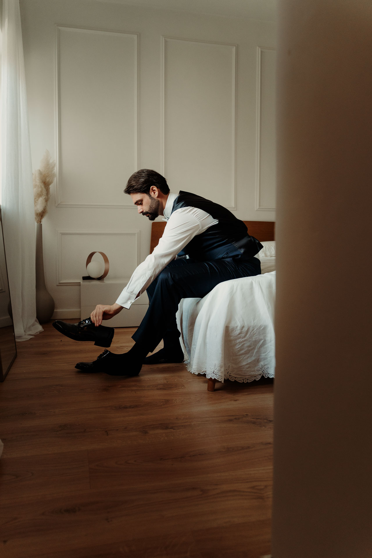 Lo sposo seduto sul letto si allaccia le scarpe, visto attraverso la porta socchiusa