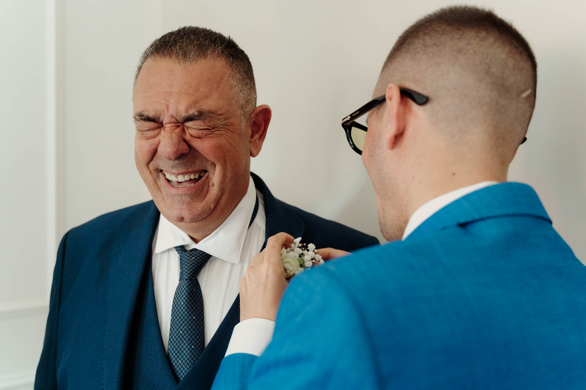 Un uomo in giacca blu ride a occhi chiusi mentre un amico con gli occhiali gli sistema il fiore all'occhiello