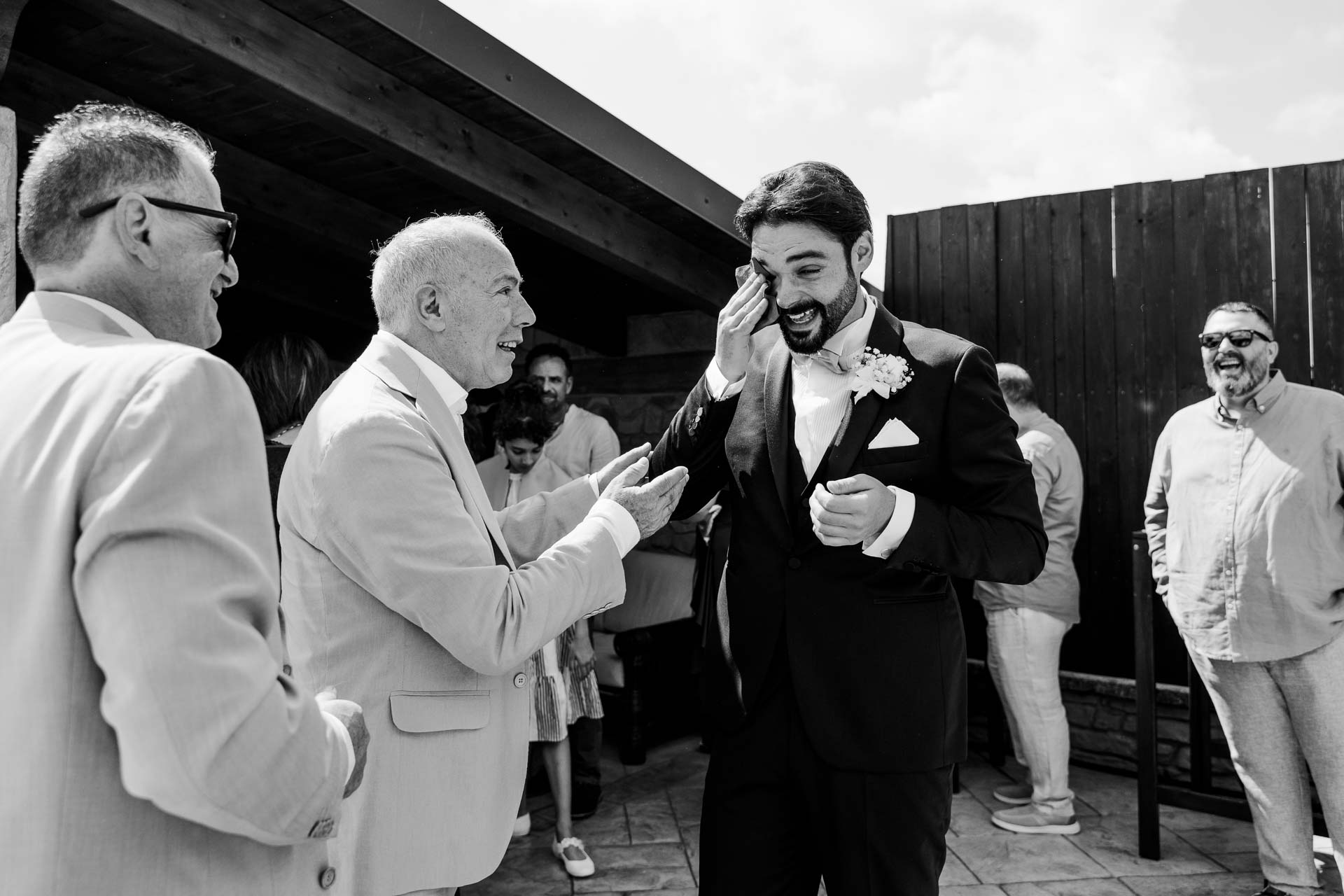 In bianco e nero, lo sposo si asciuga una lacrima mentre un invitato in giacca chiara gli parla
