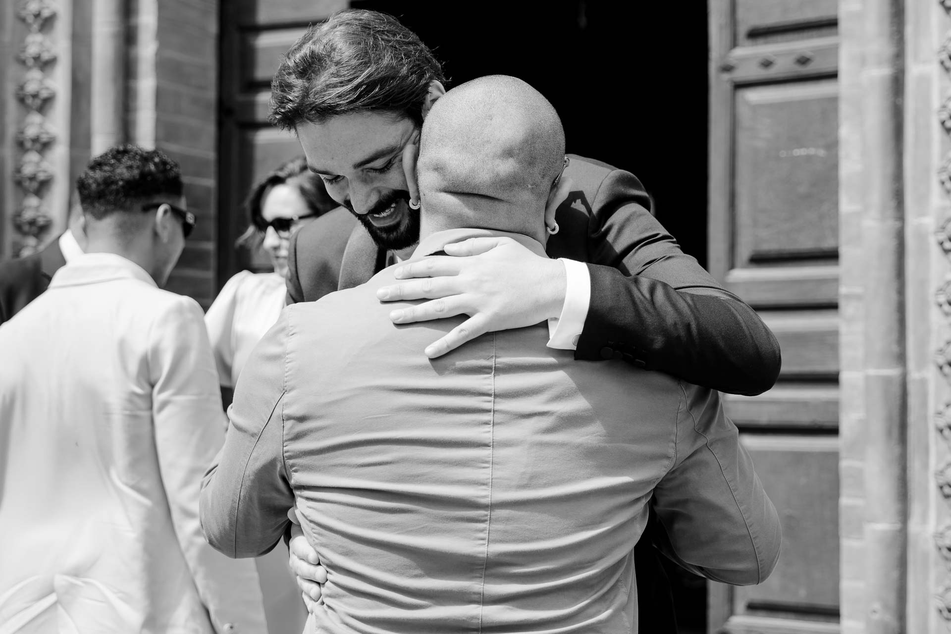 In bianco e nero, lo sposo abbraccia un amico sul sagrato, davanti al portone della chiesa