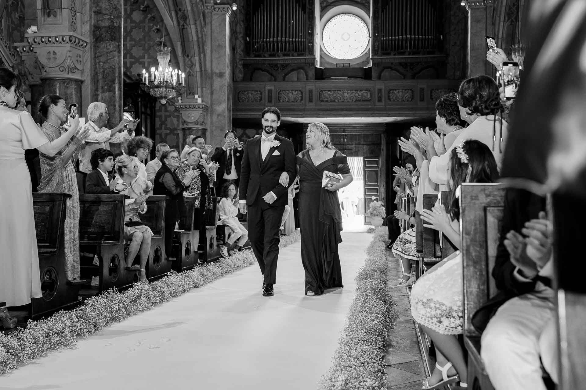 In bianco e nero, lo sposo percorre la navata sul tappeto bianco, tra gli invitati in piedi