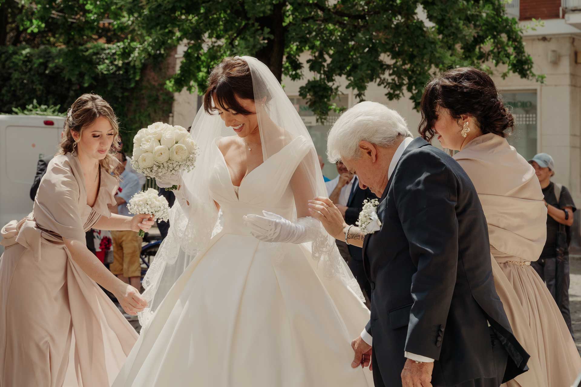 L'amica in rosa cipria con il bouquet e un signore in abito scuro aiutano la sposa a sistemare l'abito davanti alla chiesa