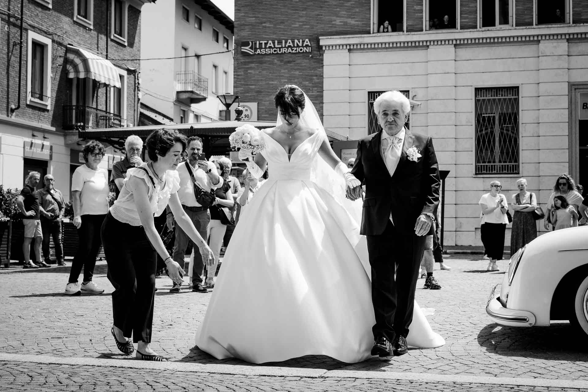 In bianco e nero, la sposa attraversa la piazza sottobraccio a un signore in abito scuro, accanto all'auto d'epoca