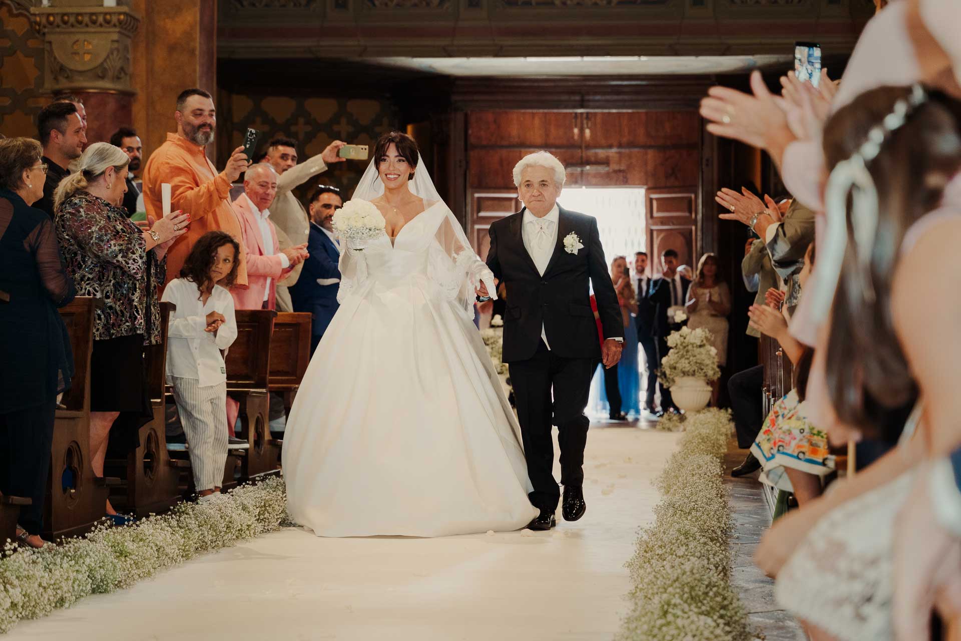 La sposa entra in chiesa a braccio di un signore in abito scuro, tra gli invitati che applaudono e filmano