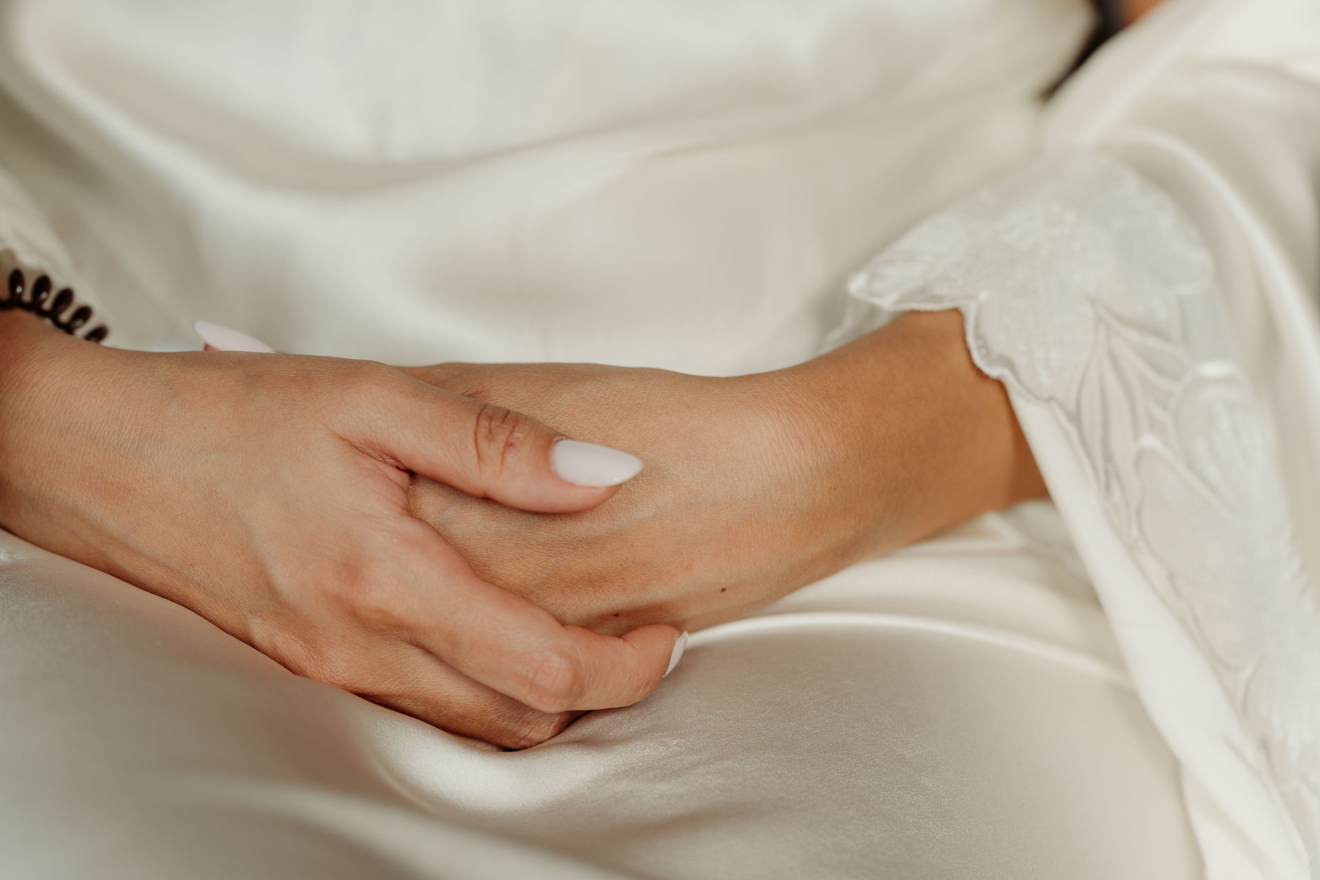 Le mani della sposa intrecciate sulla vestaglia di raso, con le maniche di pizzo
