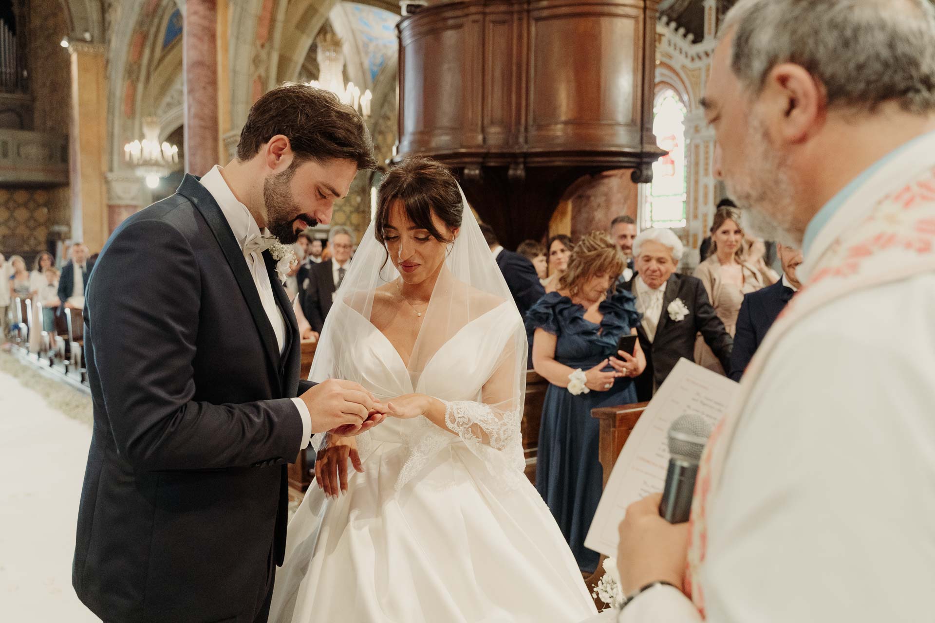 Lo sposo infila la fede alla sposa, con il sacerdote accanto