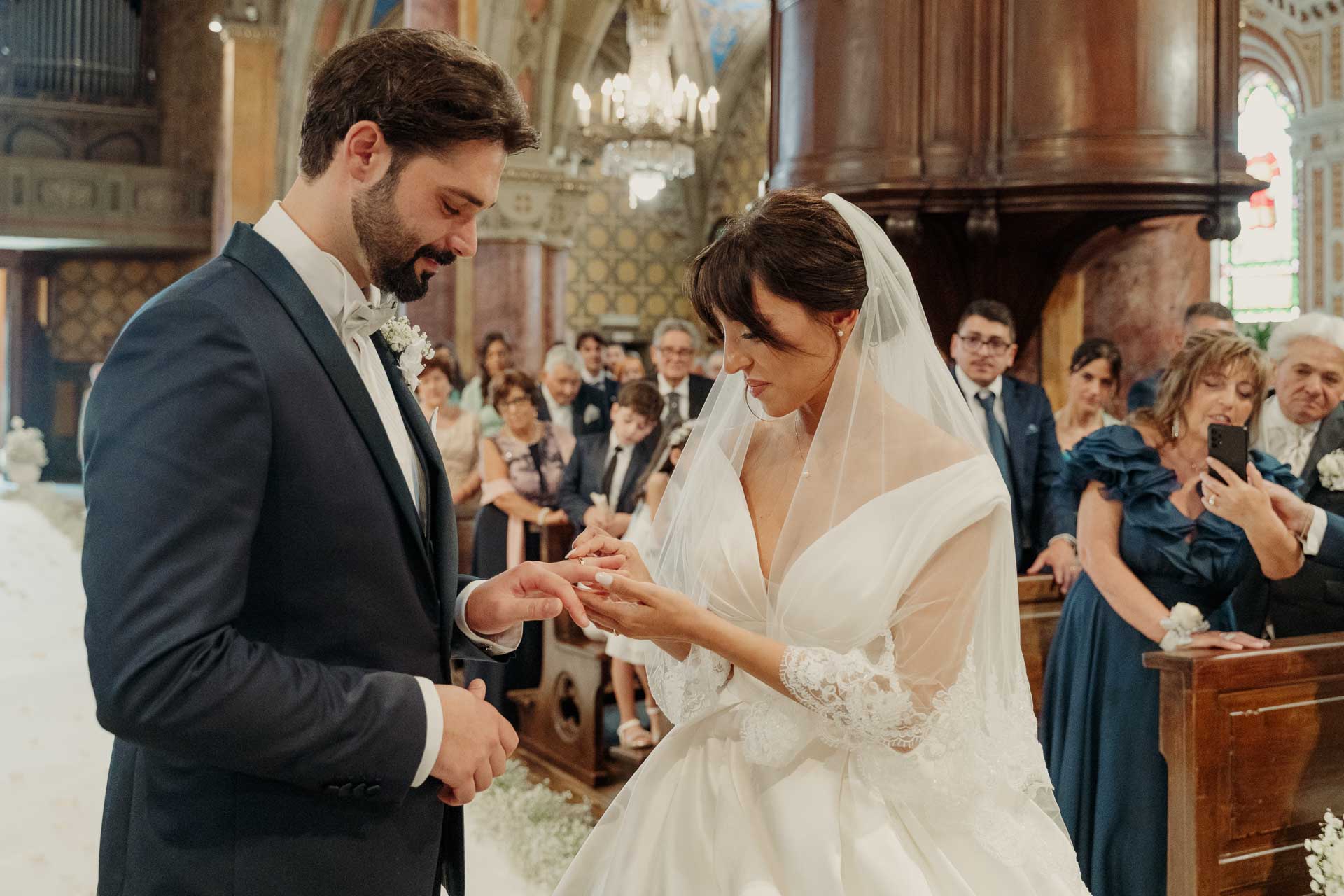 Lo sposo infila la fede alla sposa, sotto lo sguardo degli invitati
