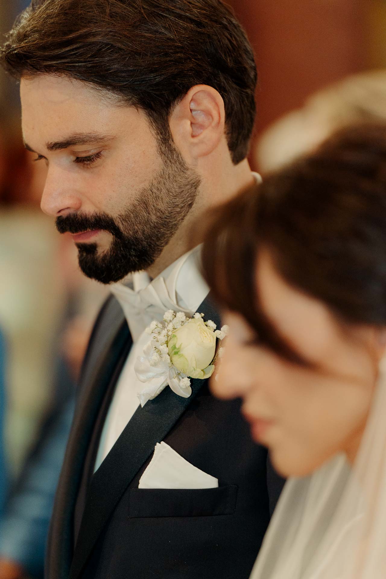 Lo sposo con lo sguardo basso accanto alla sposa, con il fiore all'occhiello di rose bianche