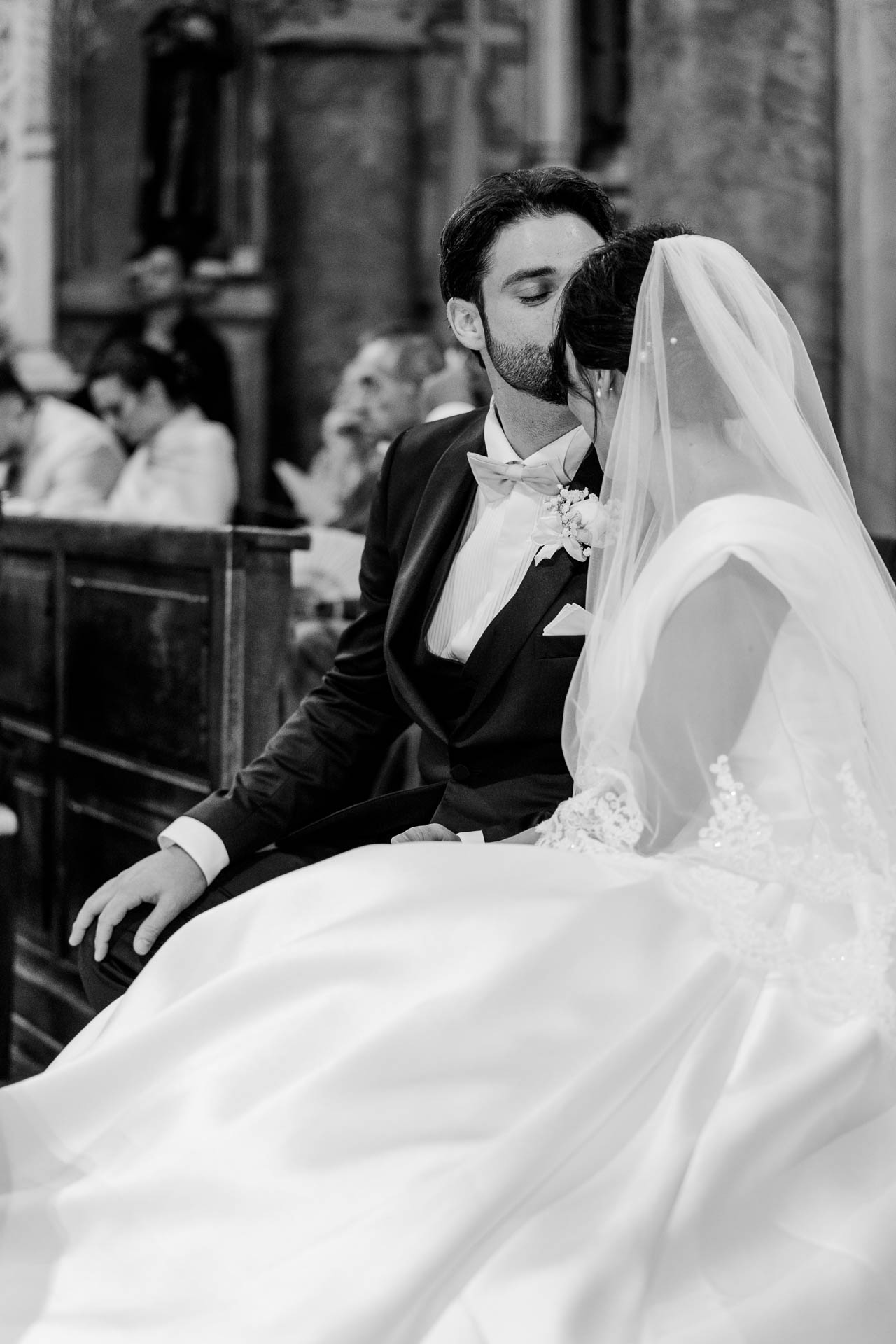 In bianco e nero, lo sposo bacia la sposa sulla tempia, seduti nel banco