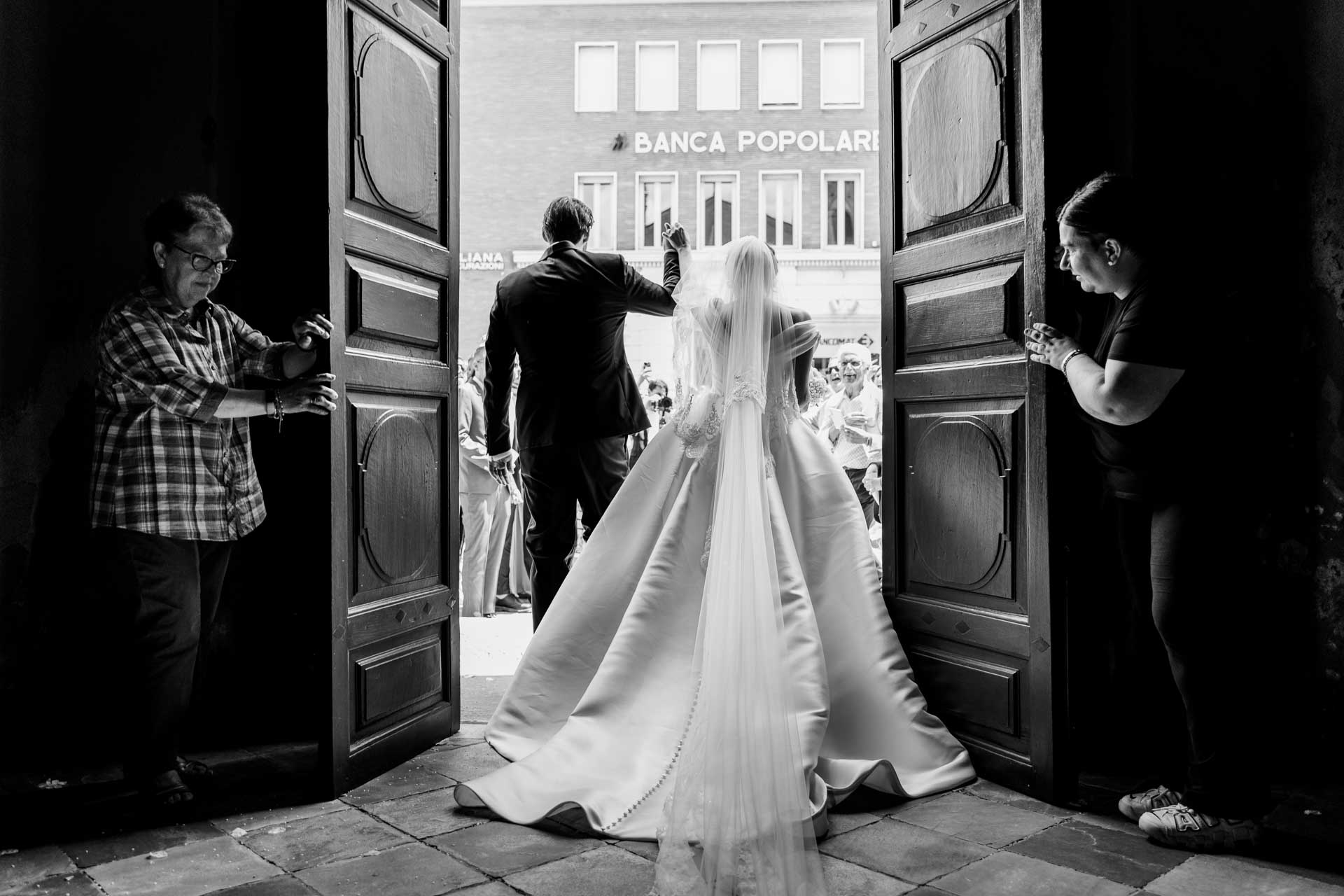 In bianco e nero, la sposa di spalle con lo strascico sulla soglia, tra i battenti aperti del portone