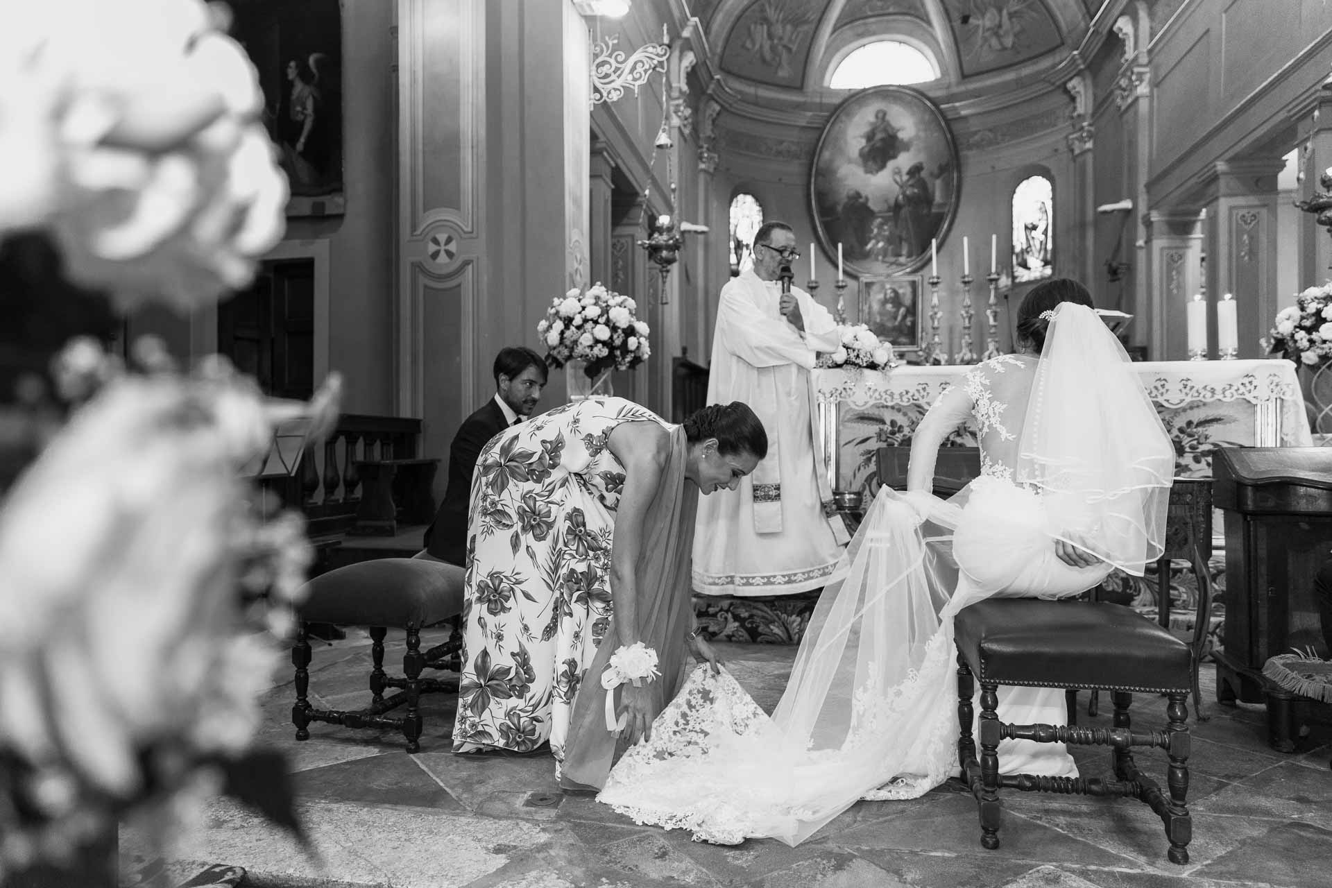 In bianco e nero, un'amica sistema lo strascico della sposa davanti all'altare
