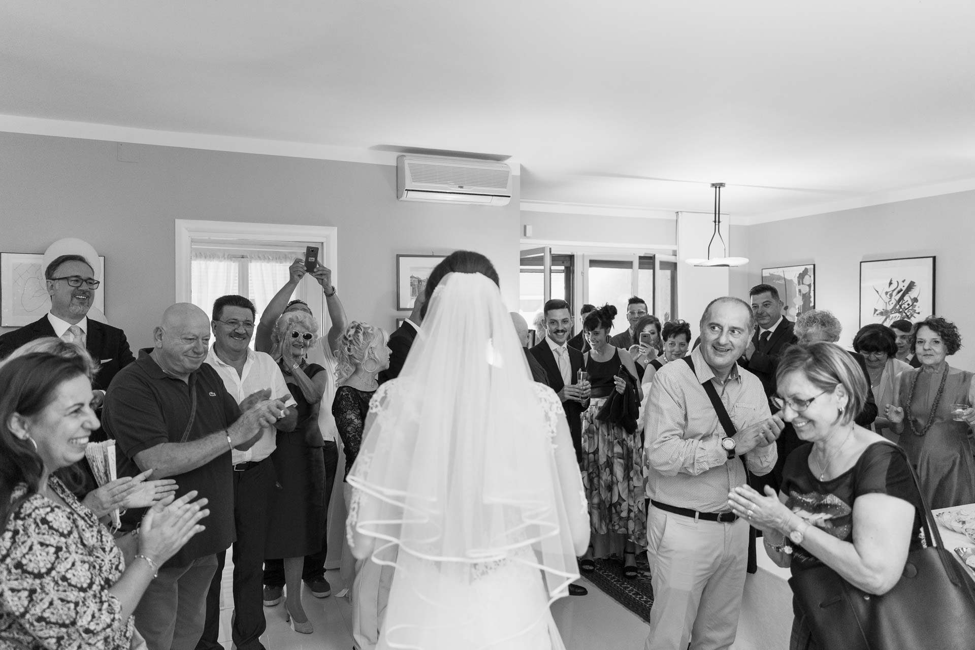 In bianco e nero, la sposa di spalle con il velo, davanti ai parenti che applaudono